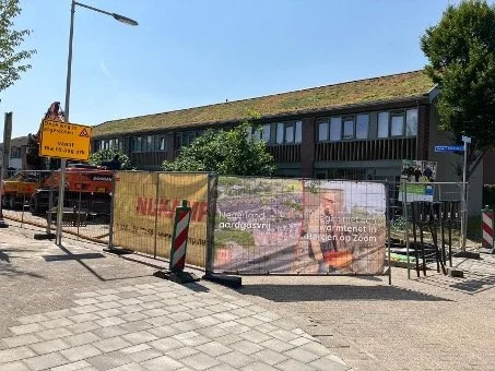 Een wegwerkersbord en hekwerken op een straat, met een gebouw en bomen op de achtergrond.