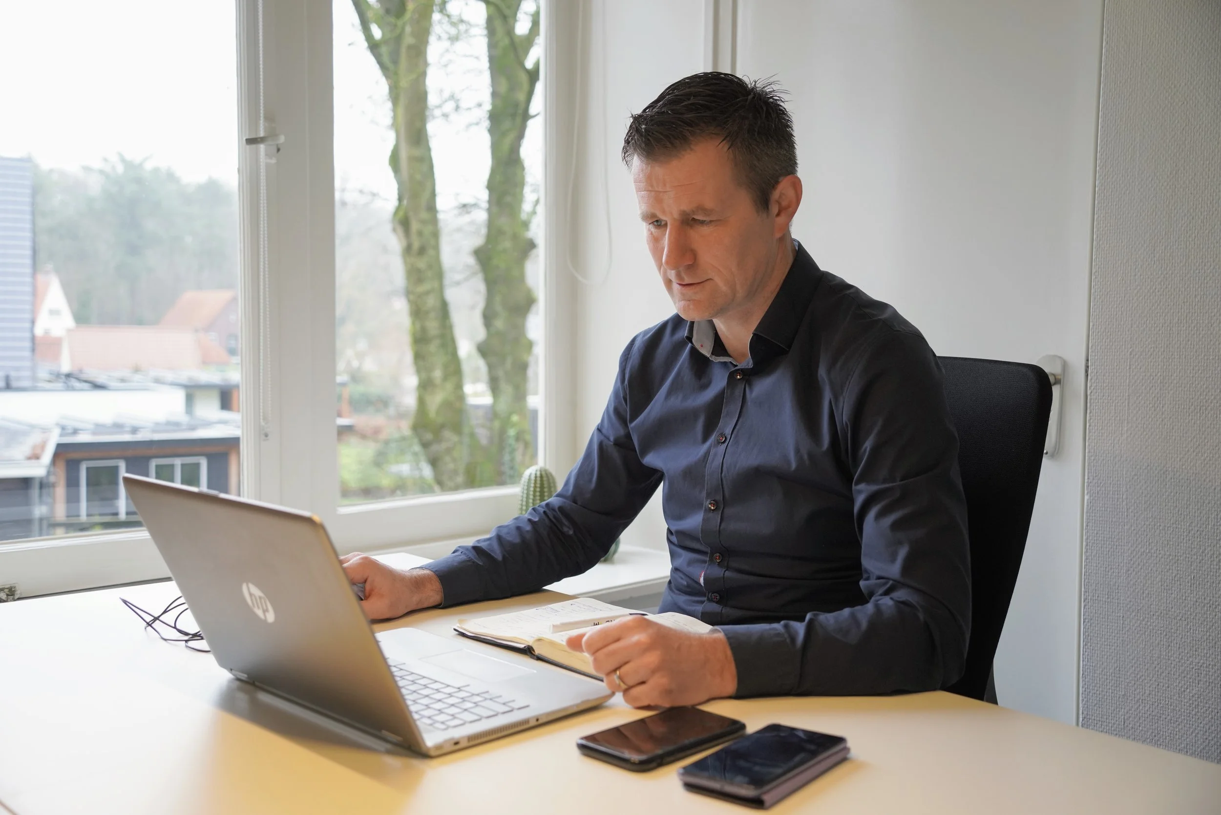 Man werkt aan laptop aan bureau met notitieboekje en smartphones in kantoor met raam en uitzicht op bomen buiten.