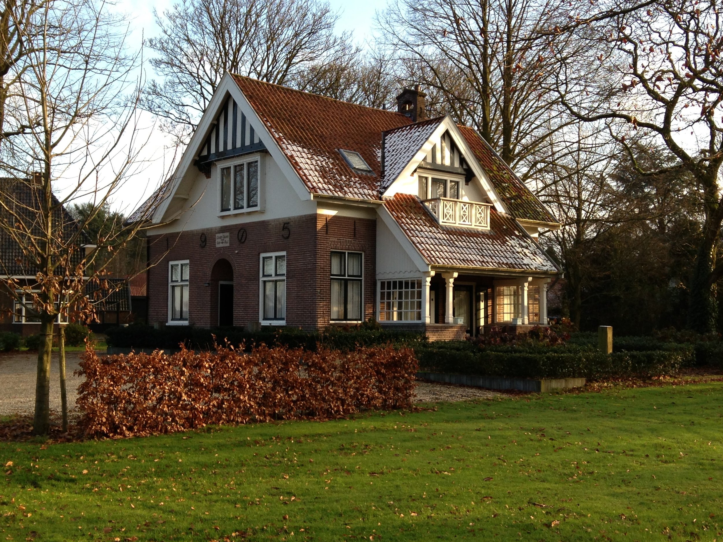 Een grote, oude huis met een rood dak en witte gevels, omringd door bomen en een tuin met gras en struiken.