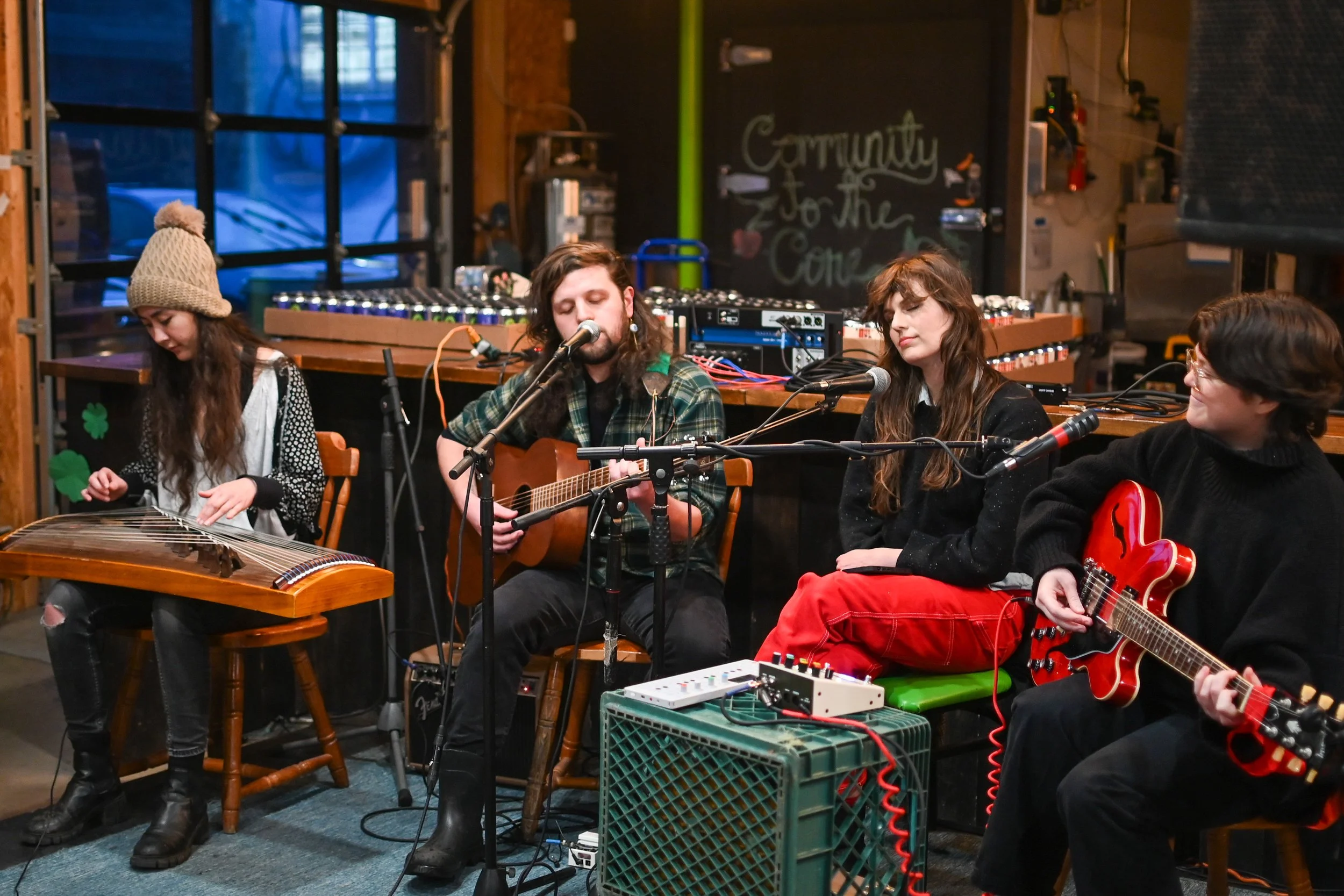 Four musicians performing in a cozy indoor setting. A woman wearing a gray knit hat and black boots is playing a hammered dulcimer, a man in the center is singing and playing an acoustic guitar, a woman next to him is singing, and a woman on the right is playing an electric guitar. There is a chalkboard in the background with writing that says 'Community to the Core'.