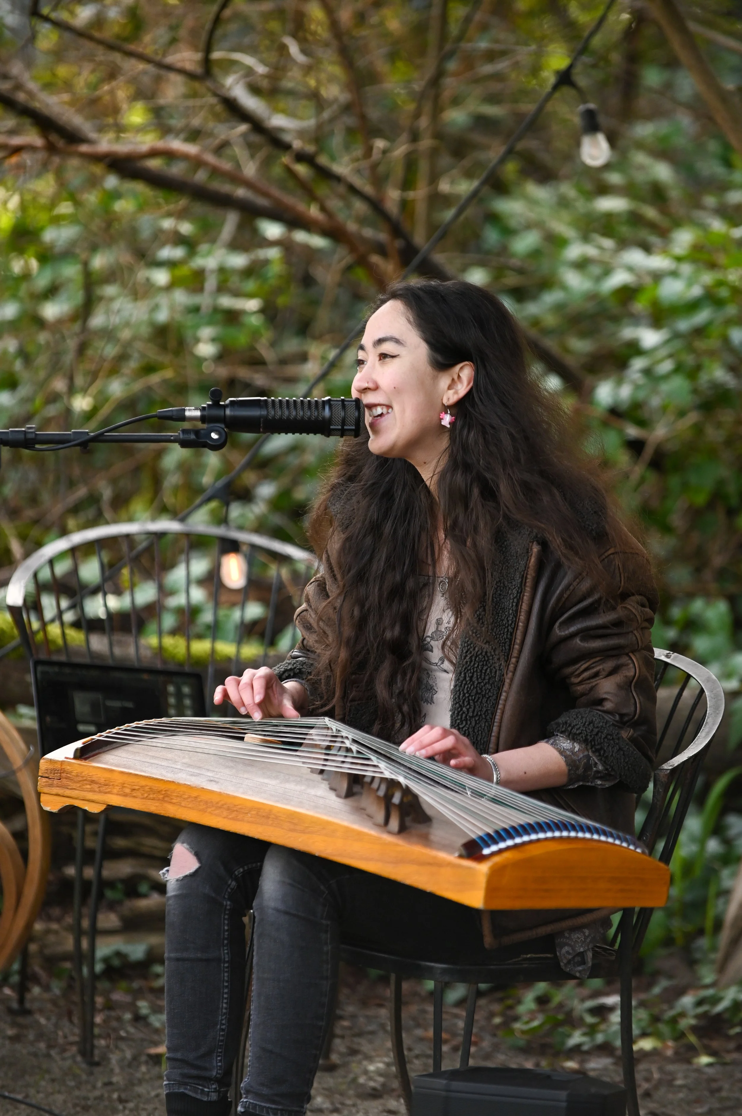 A woman with long dark hair, wearing a leather jacket and patterned top, is playing a hammered dulcimer outdoors while smiling and singing into a microphone.