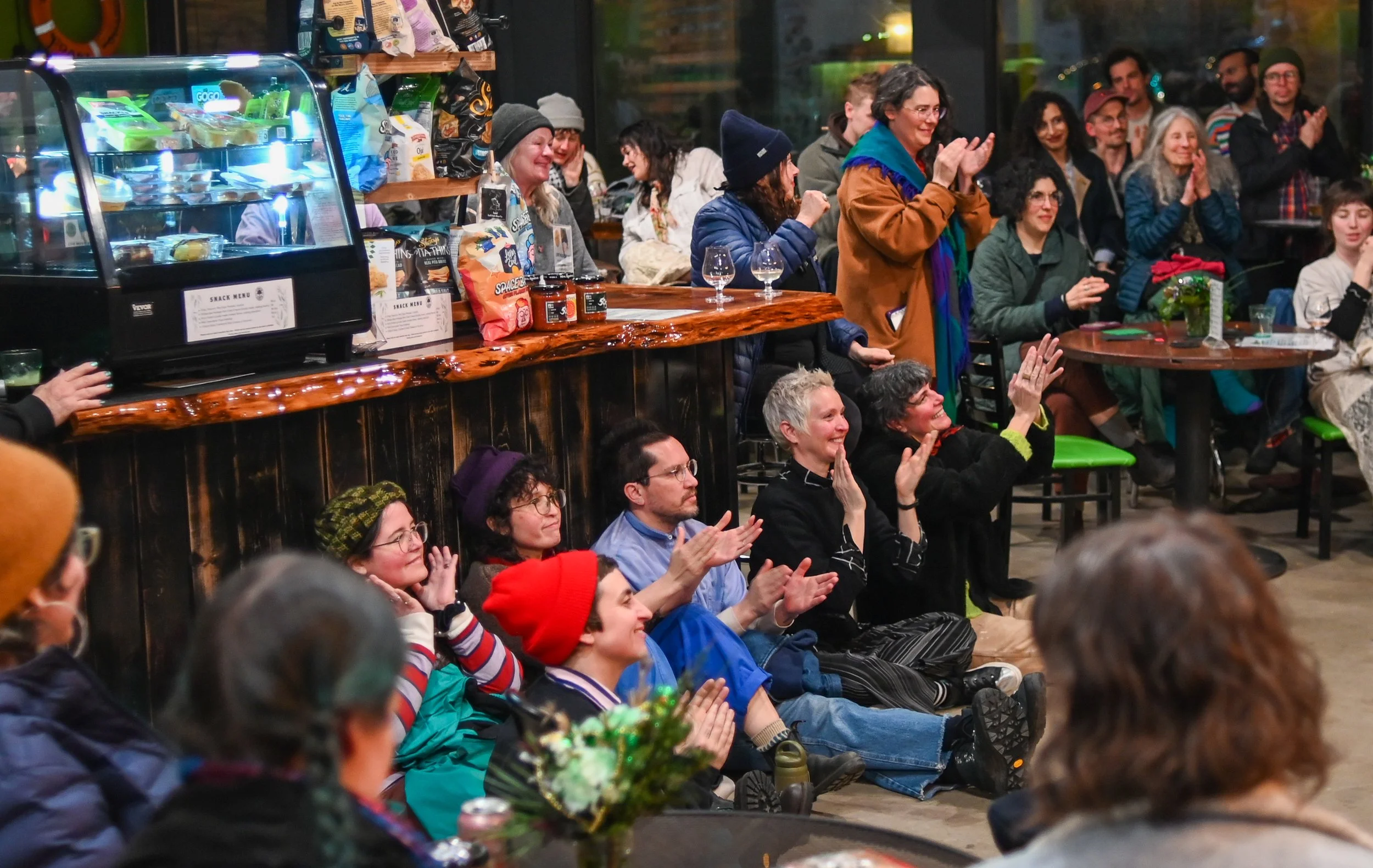 People gathered in a cozy, indoor space, most seated on the floor and chairs, watching and clapping during a live event or performance. Some are seated at a bar with snacks and drinks, with a glass display case containing snacks visible on the bar. The background shows a window with a view of the outdoors, and the atmosphere is warm and lively.