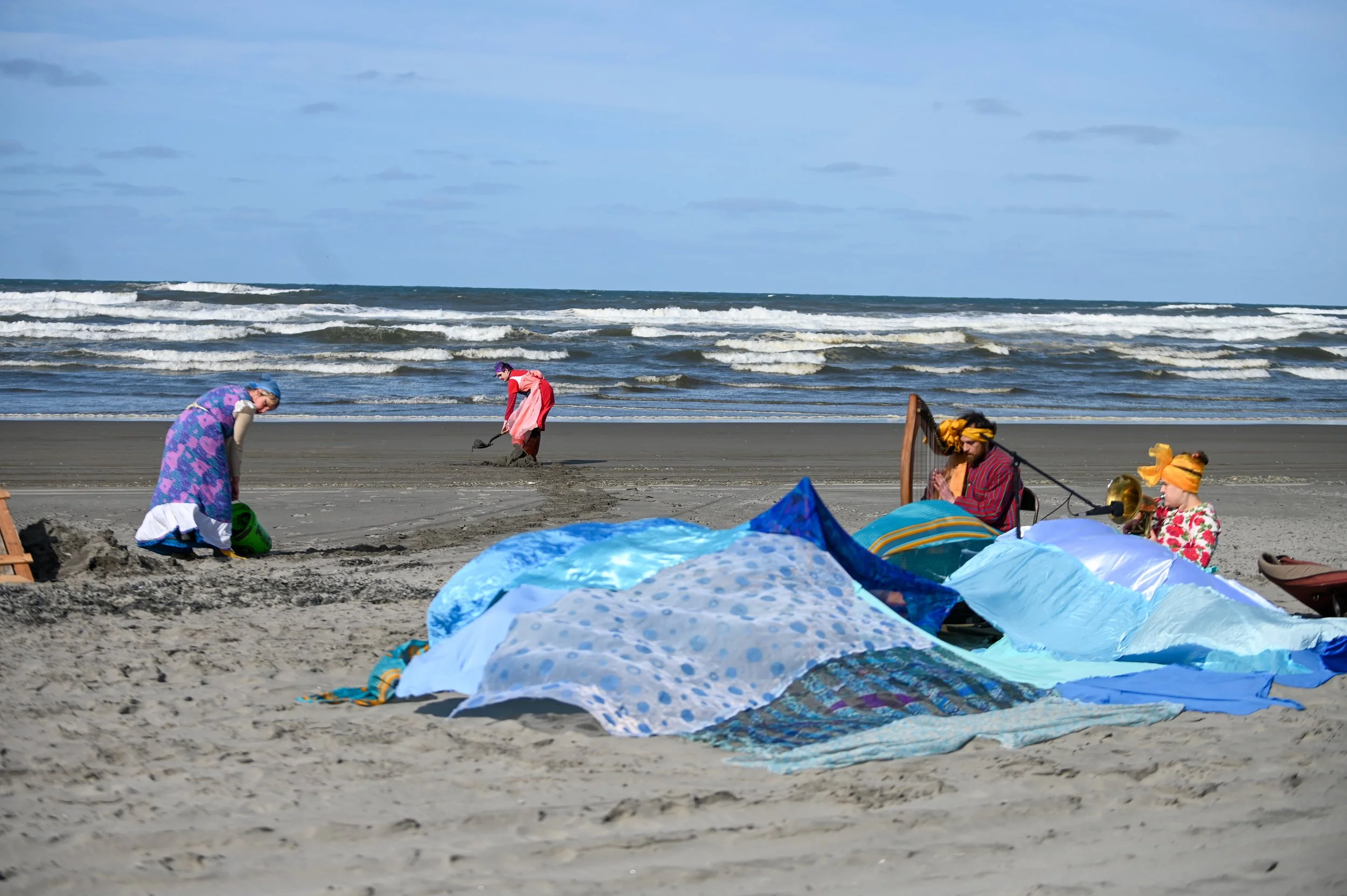 People dressed in colorful clothing and turbans on a beach, surrounded by folded umbrellas or fabrics with ocean waves in the background.
