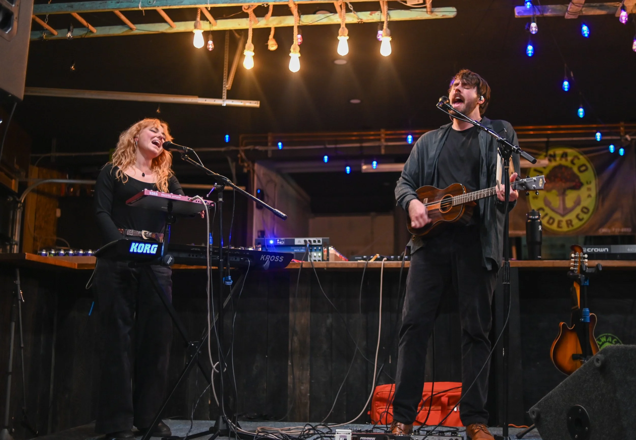 A man and woman performing on stage with musical instruments, including a keyboard and a guitar, under hanging lights with an audience and a bartender bar in the background