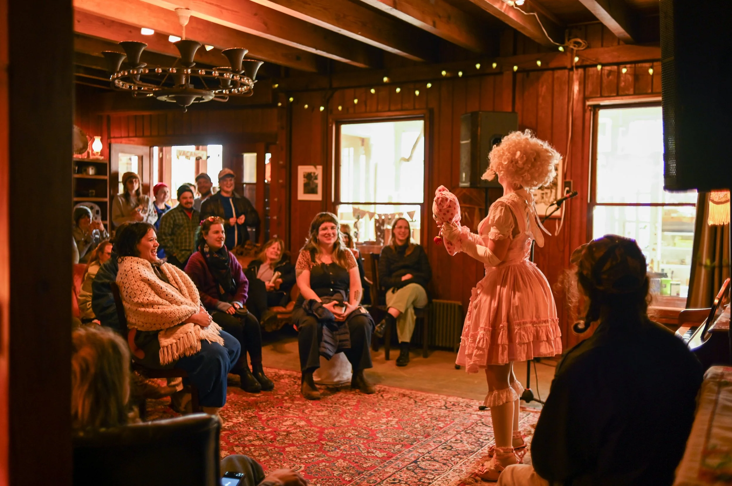 A person dressed as a clown with curly blond hair, wearing a pink dress and holding a puppet, entertains an audience in a cozy, rustic room with wood-paneled walls and string lights. The audience, consisting of various ages, is seated on chairs and benches, smiling and laughing.