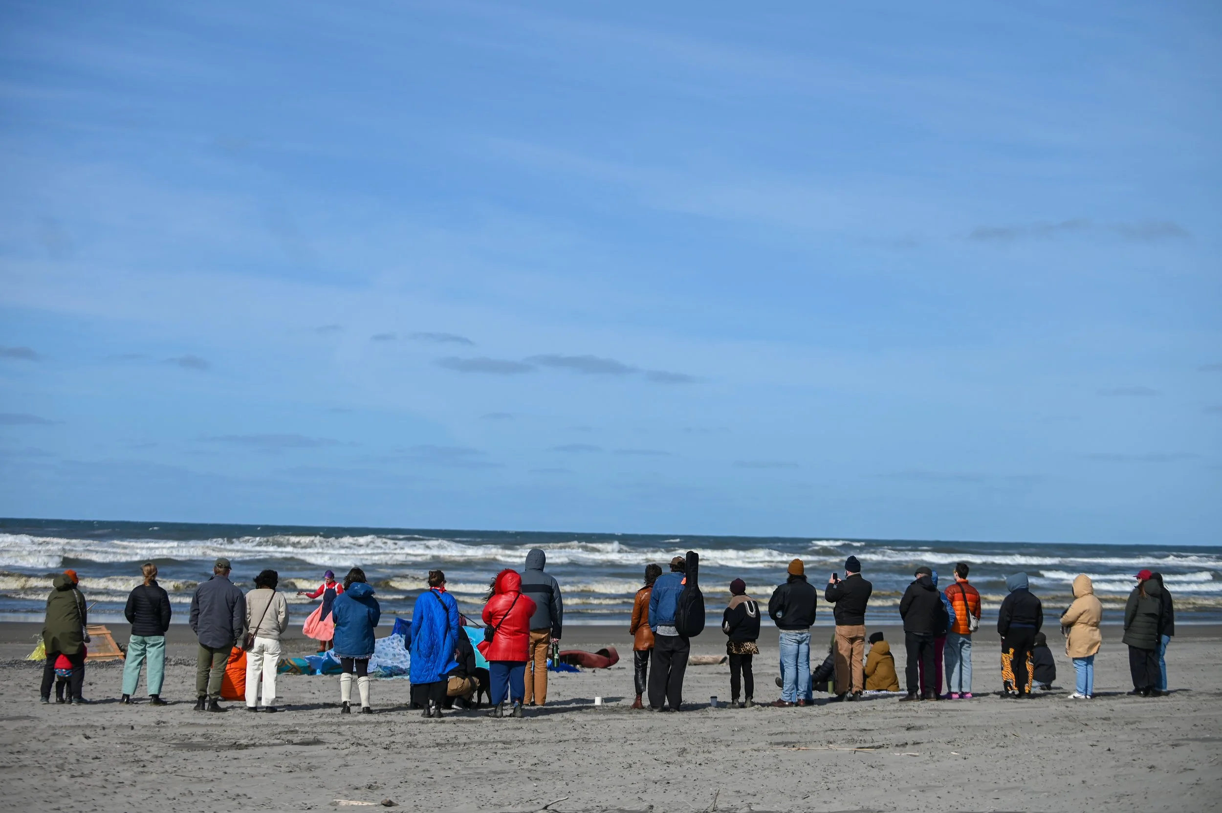 A group of people dressed in warm clothing on a sandy beach, facing the ocean with waves, under a partly cloudy sky.
