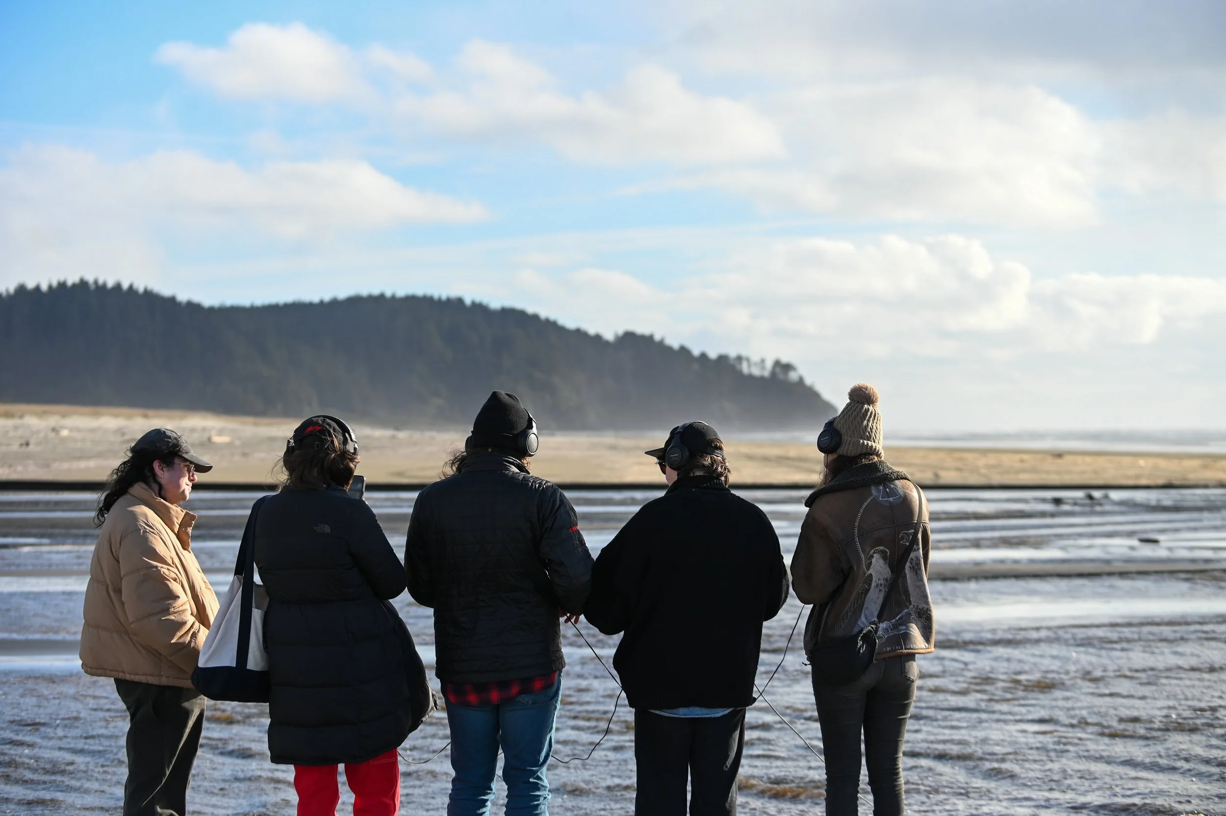 Five people standing on a beach wearing headphones, facing the ocean with a distant forested hill in the background.