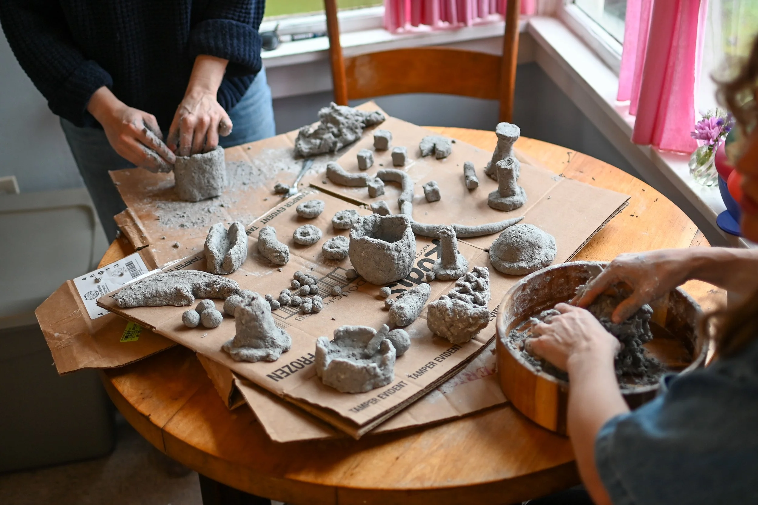 Two people creating and assembling clay or cement sculptures and figurines on a wooden table with various shaped objects, surrounded by pink curtains and sunlight.