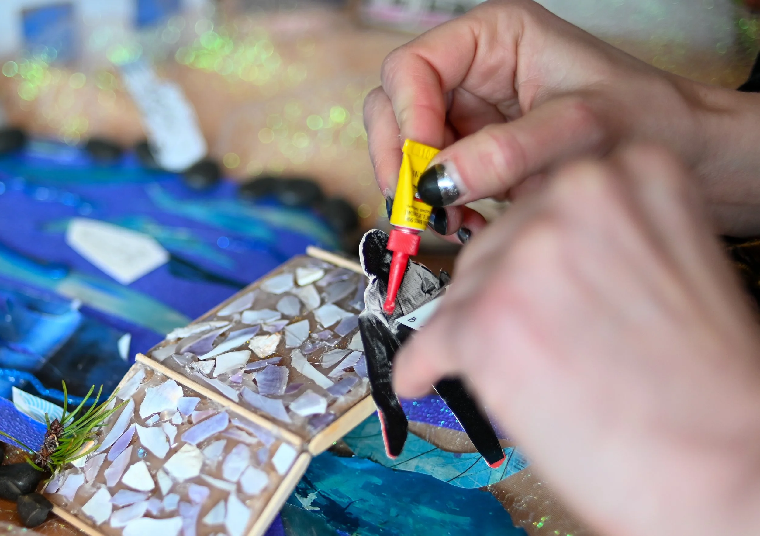 A person using a red glue tube to attach a black paper shape to a collage of seashells and stones. Blue and gold glitter and other art supplies are visible in the background.