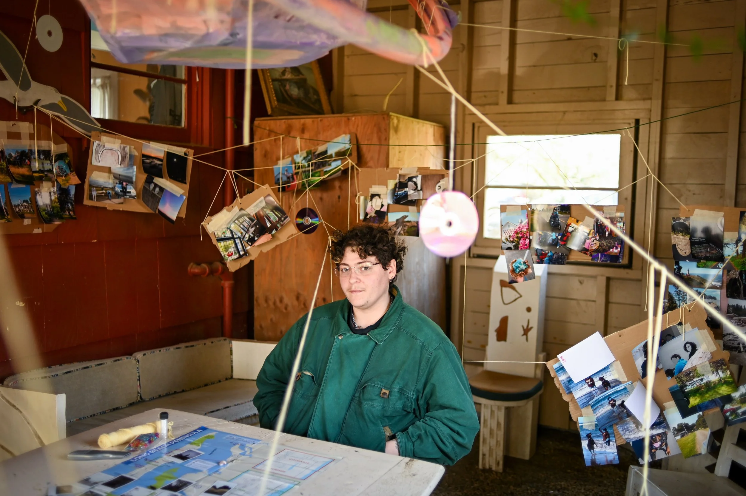 A person with short curly hair and glasses wearing a green jacket sits at a table in a room with wooden-paneled walls. The room is decorated with hanging strings of photographs and paper disks, creating a display. There are windows letting in natural light and various items on the table, including papers and a spool.