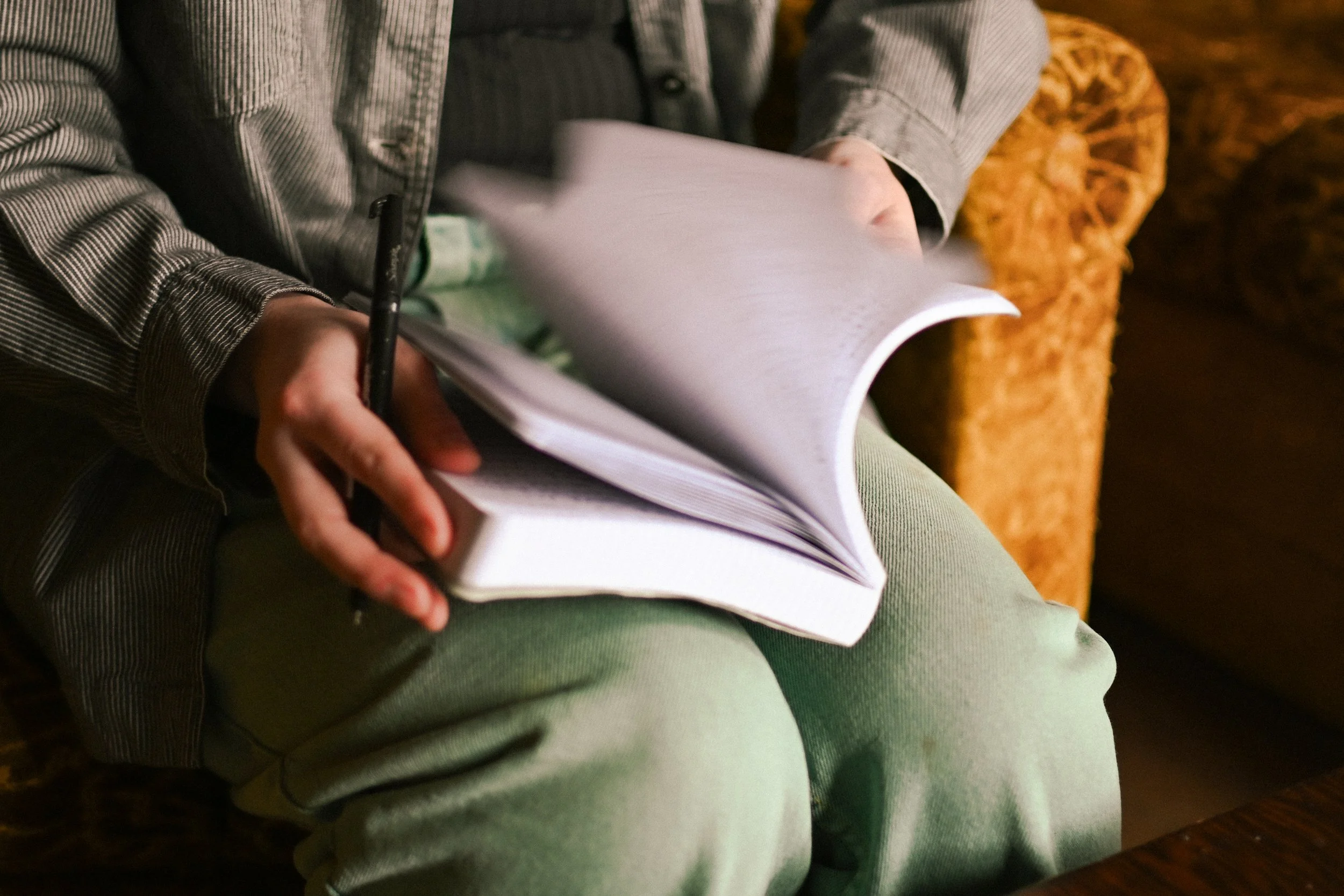 Person in a striped shirt and light green pants sitting on a wooden chair, flipping through a thick notebook or binder with loose papers inside.