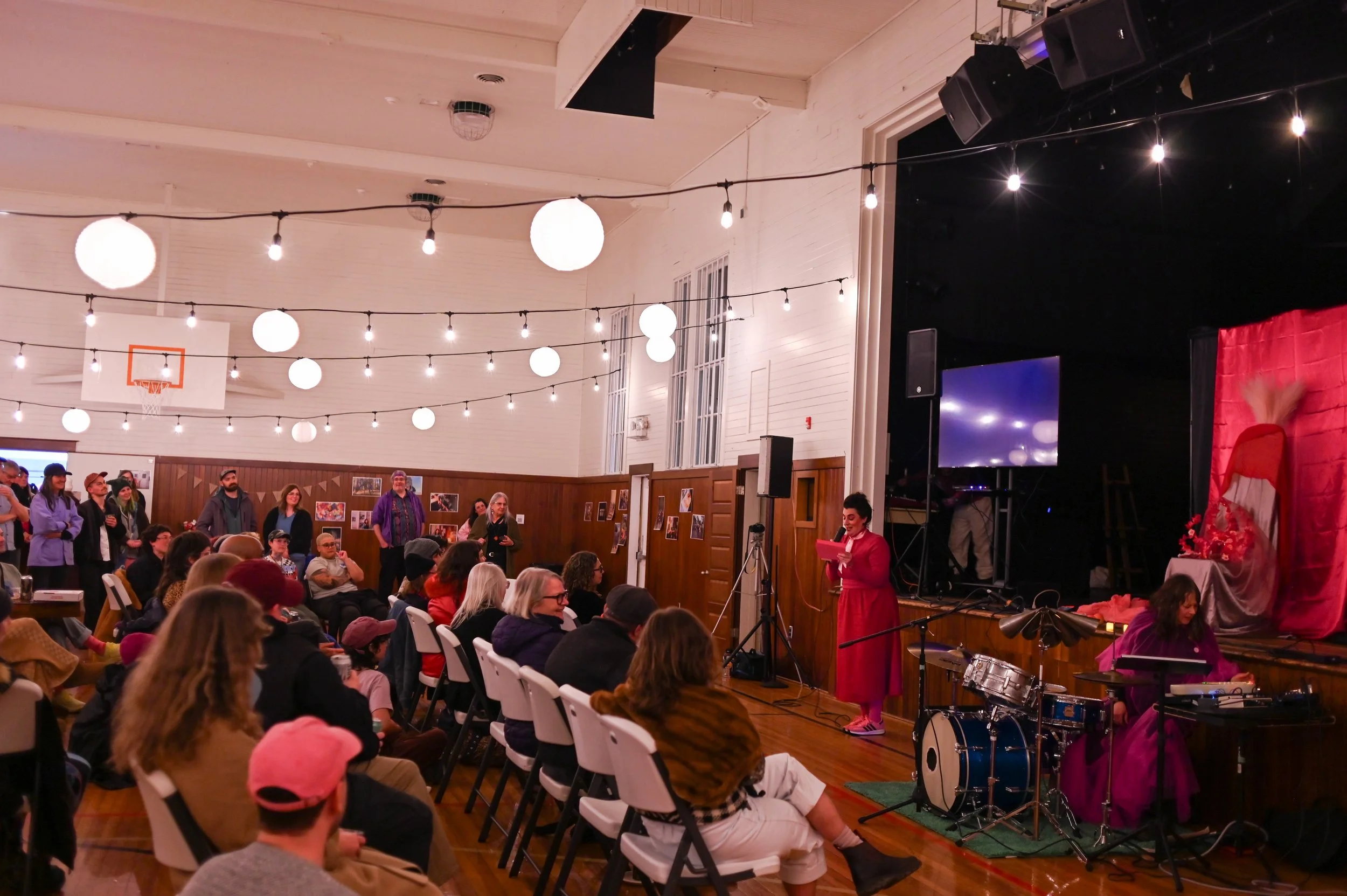 Indoor event with seated audience and performers on stage, decorated with red backdrop and floral arrangements, string lights hanging from the ceiling, and basketball hoop visible in the background.