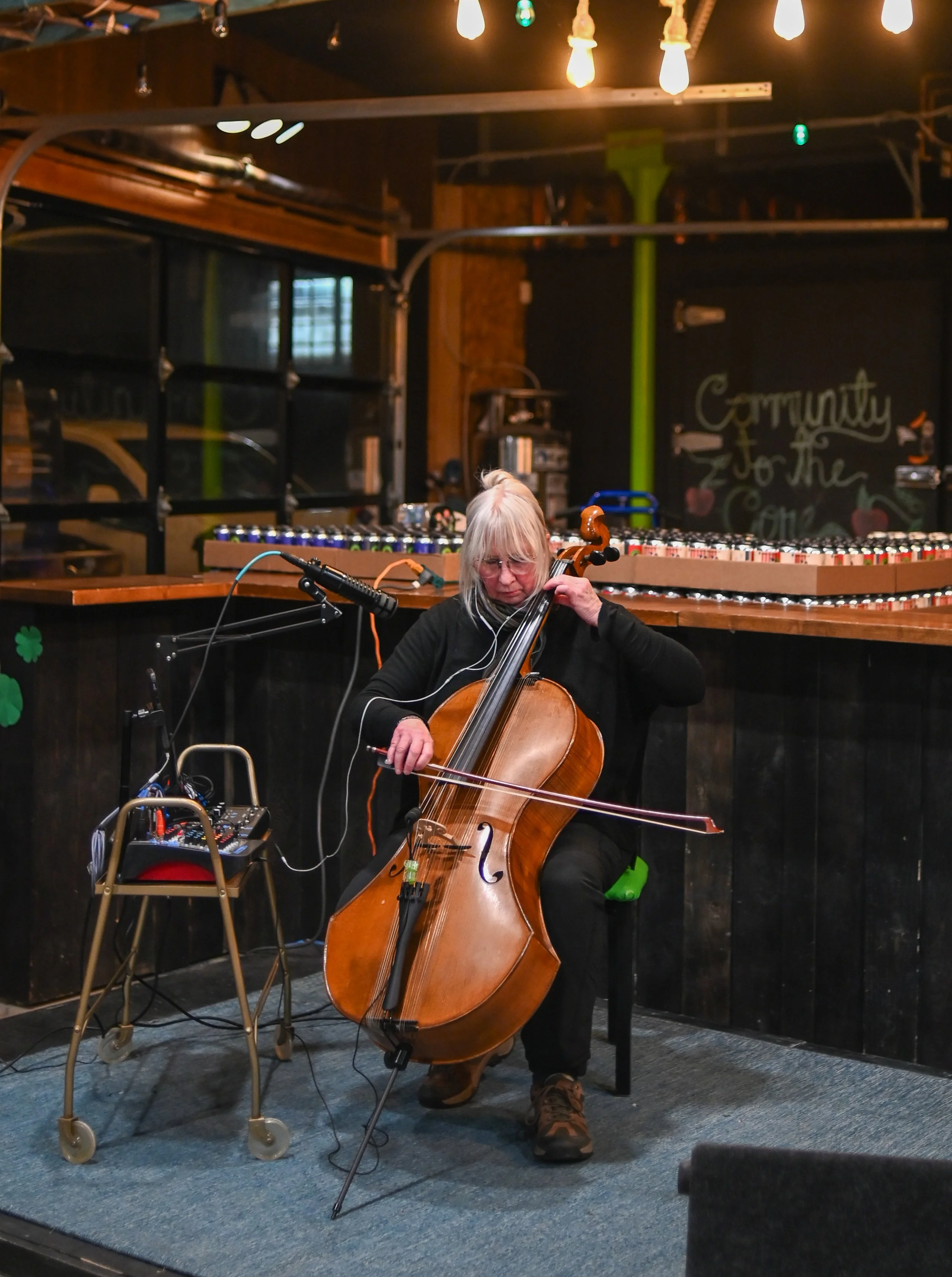 A woman with gray hair, glasses, and black clothing playing a cello in a cozy, dimly-lit bar or cafe with warm string lights.