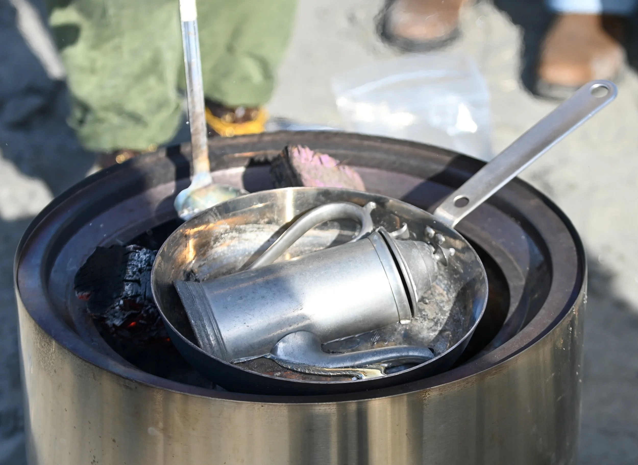 Empty beverage can and metal cup resting on a portable camping stove with ashes and charcoal underneath.