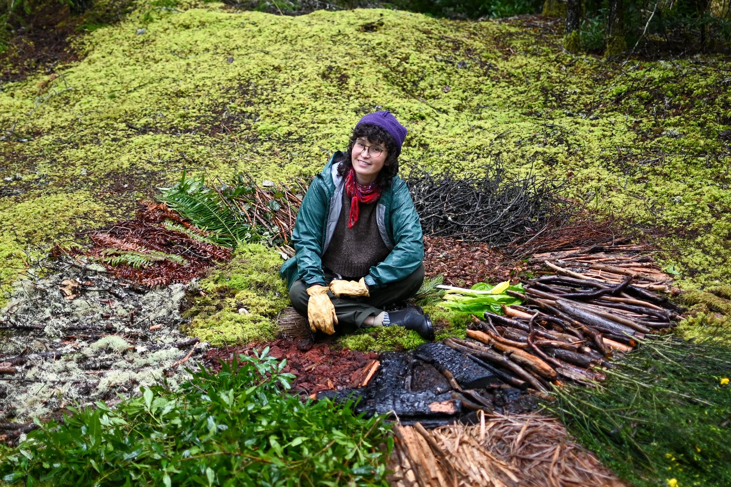 Person sitting on the ground in a forest, surrounded by plants and moss, with collected plant materials or firewood arranged around them, wearing outdoor clothing and gloves.