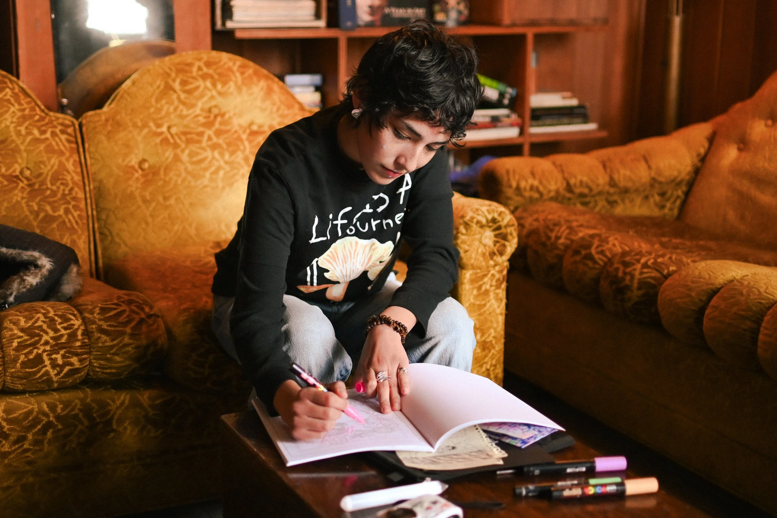 A young person with short, curly dark hair sitting on a brown, patterned sofa, writing in a notebook placed on a wooden coffee table, with various markers and art supplies nearby, in a cozy room with wooden bookshelves and warm lighting.