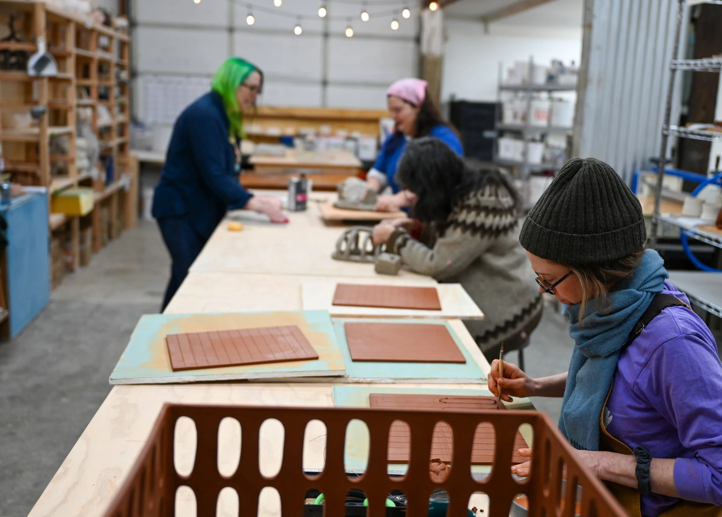 People working on woodworking projects at a long worktable in a workshop, with shelves of tools and materials in the background.