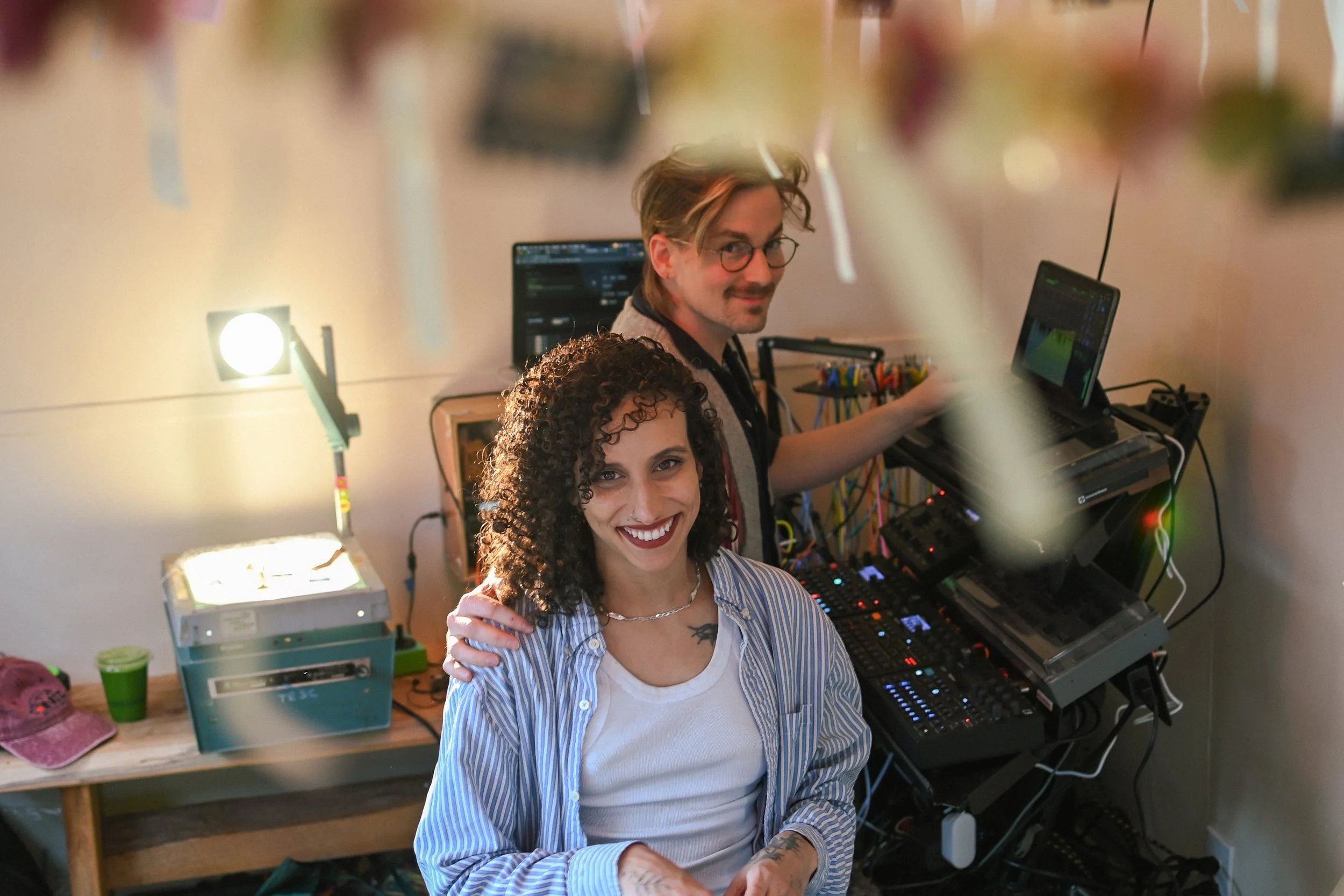 A woman with curly hair and a man with glasses and light hair smiling in a DJ or music setup room, with electronic equipment, computers, and colorful controls around them.