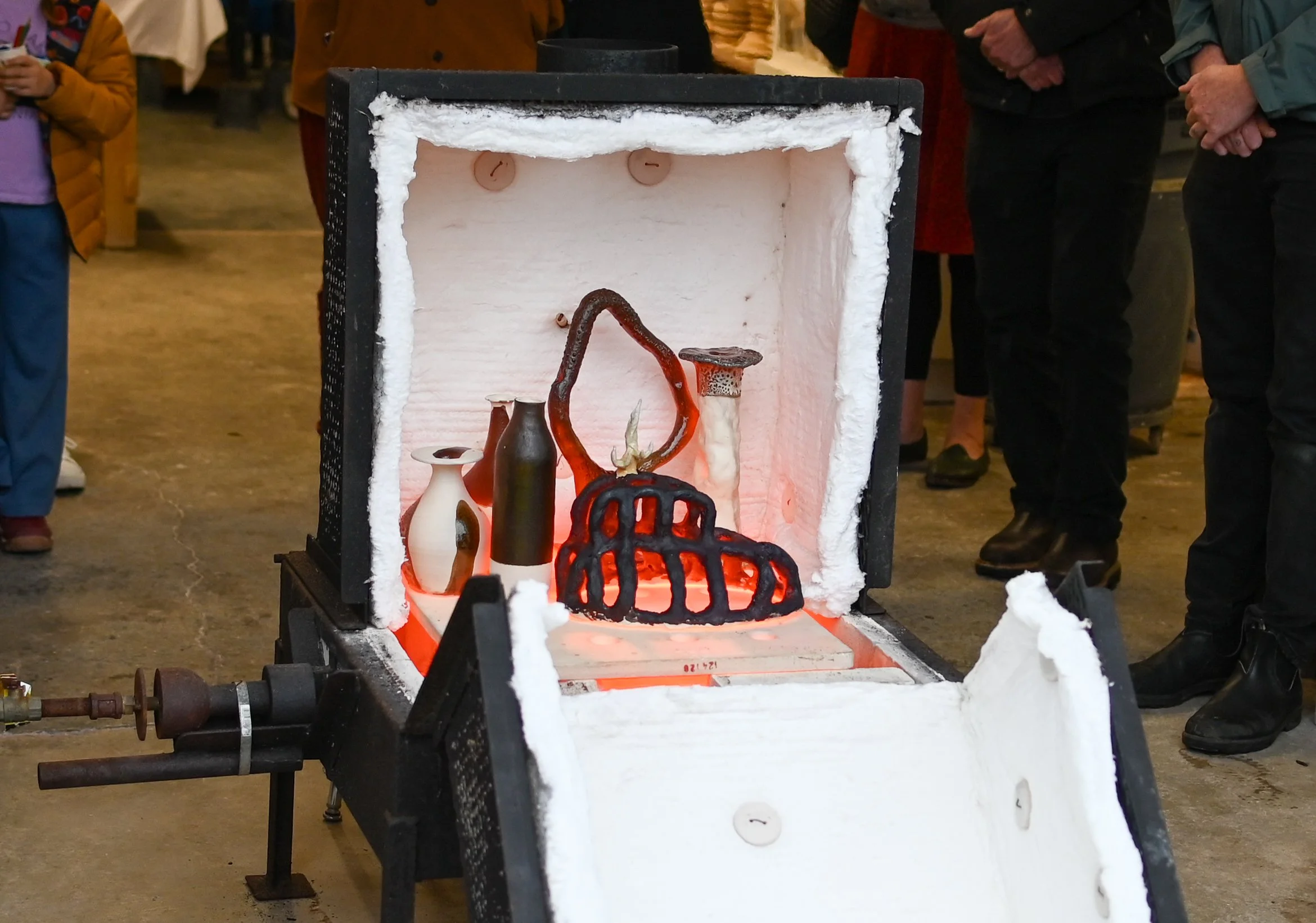 A glass-blowing furnace with a glowing red hot glass object inside, surrounded by cooling insulation, with people observing in the background.
