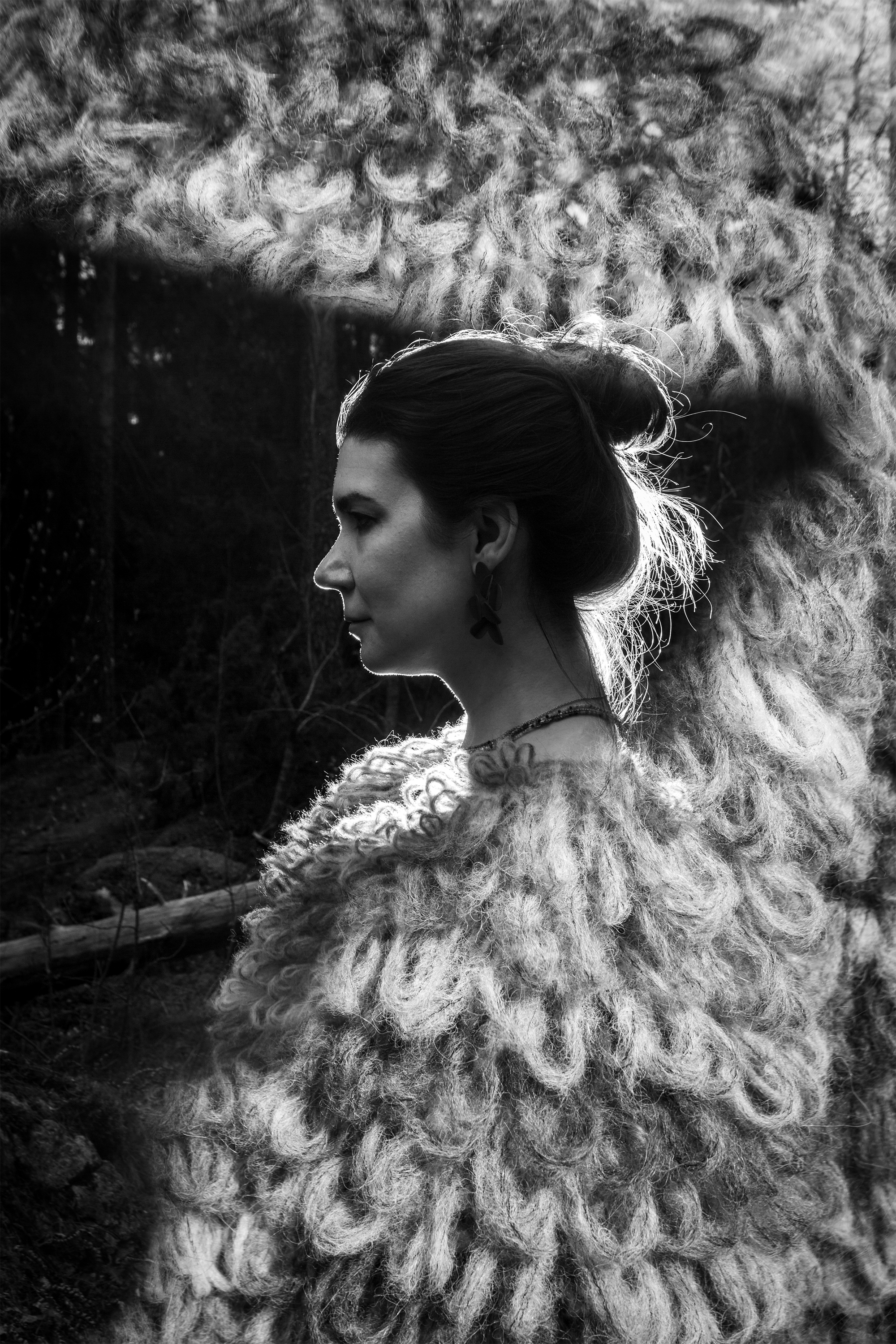 Side profile of a woman with dark hair tied back, wearing earrings and a fur coat, standing outdoors in a natural setting.