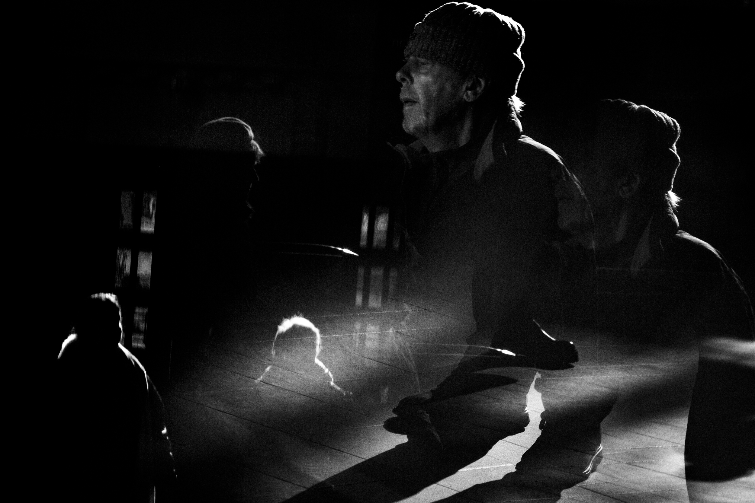 Black and white photo of multiple people, some visible in shadow and silhouette, with intense lighting on their faces, gathered in a dimly lit space.