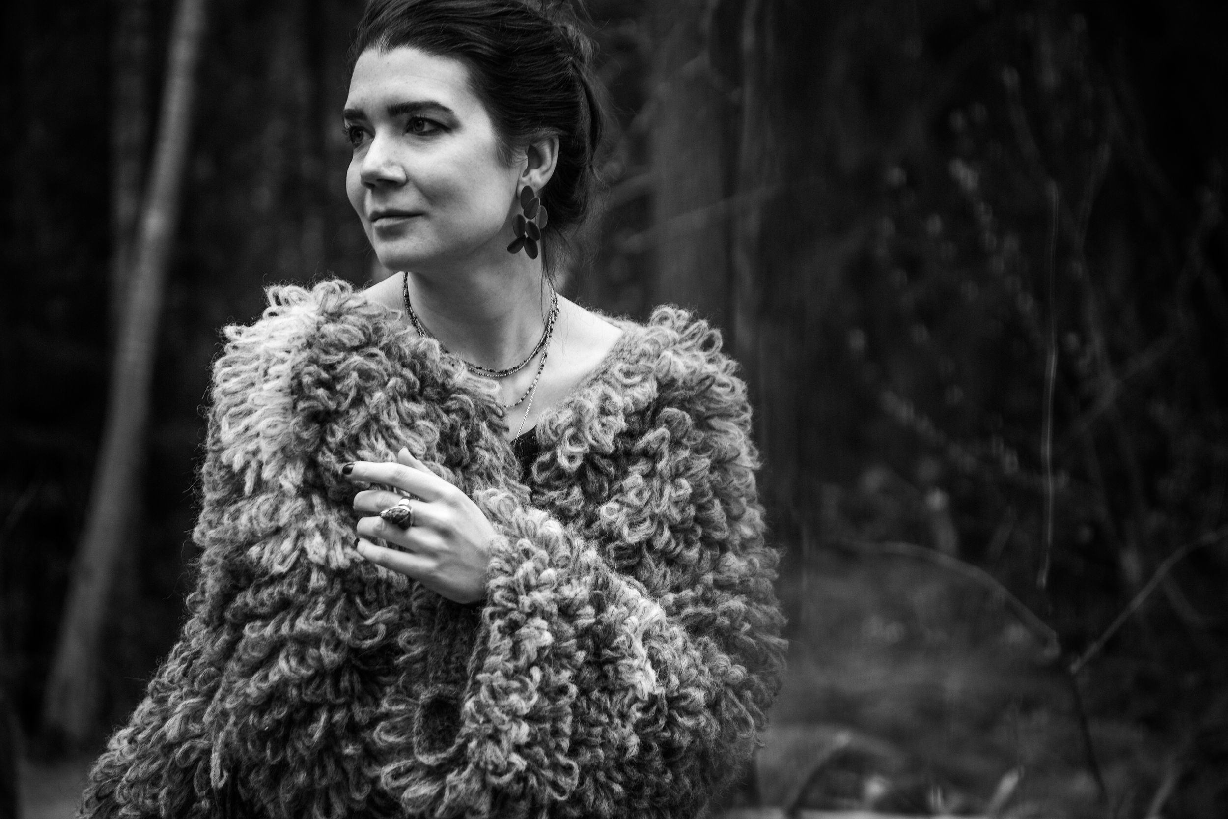 A woman with dark hair styled up, wearing patterned earrings, layered necklaces, and a textured, curly fur coat, standing outdoors in front of a blurred background of trees and foliage.