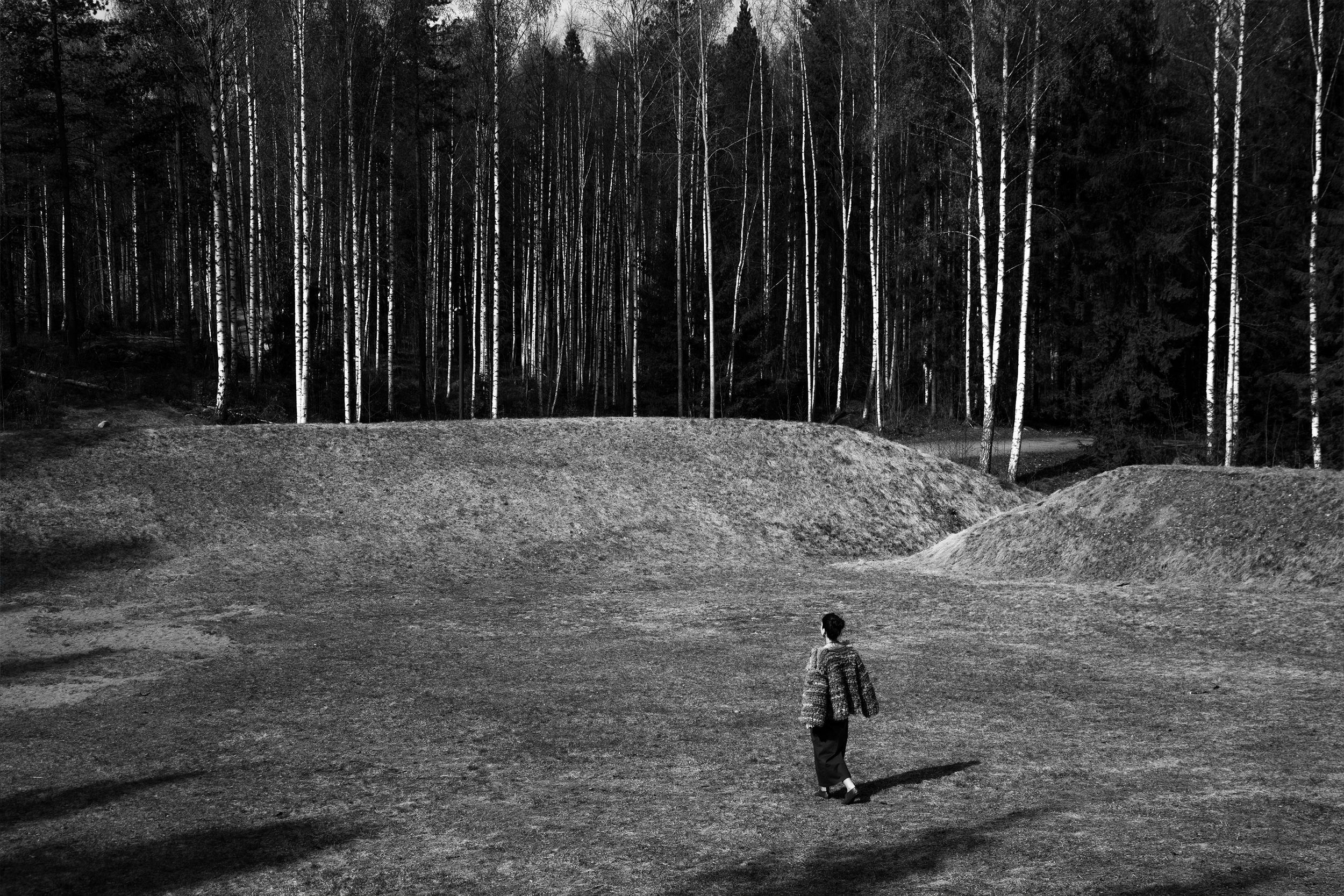 A person standing on a dirt clearing in front of a forest of tall, narrow trees with bare branches, during daytime.