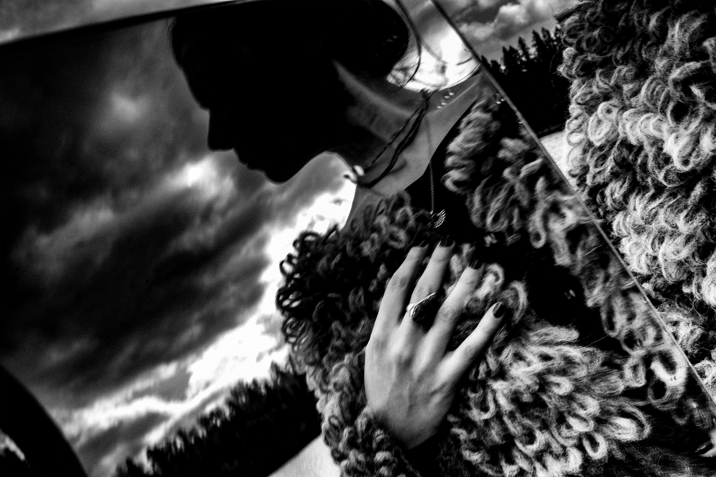Black and white photograph of a woman with curly hair resting her hand on a textured surface, with a cloudy sky in the background.