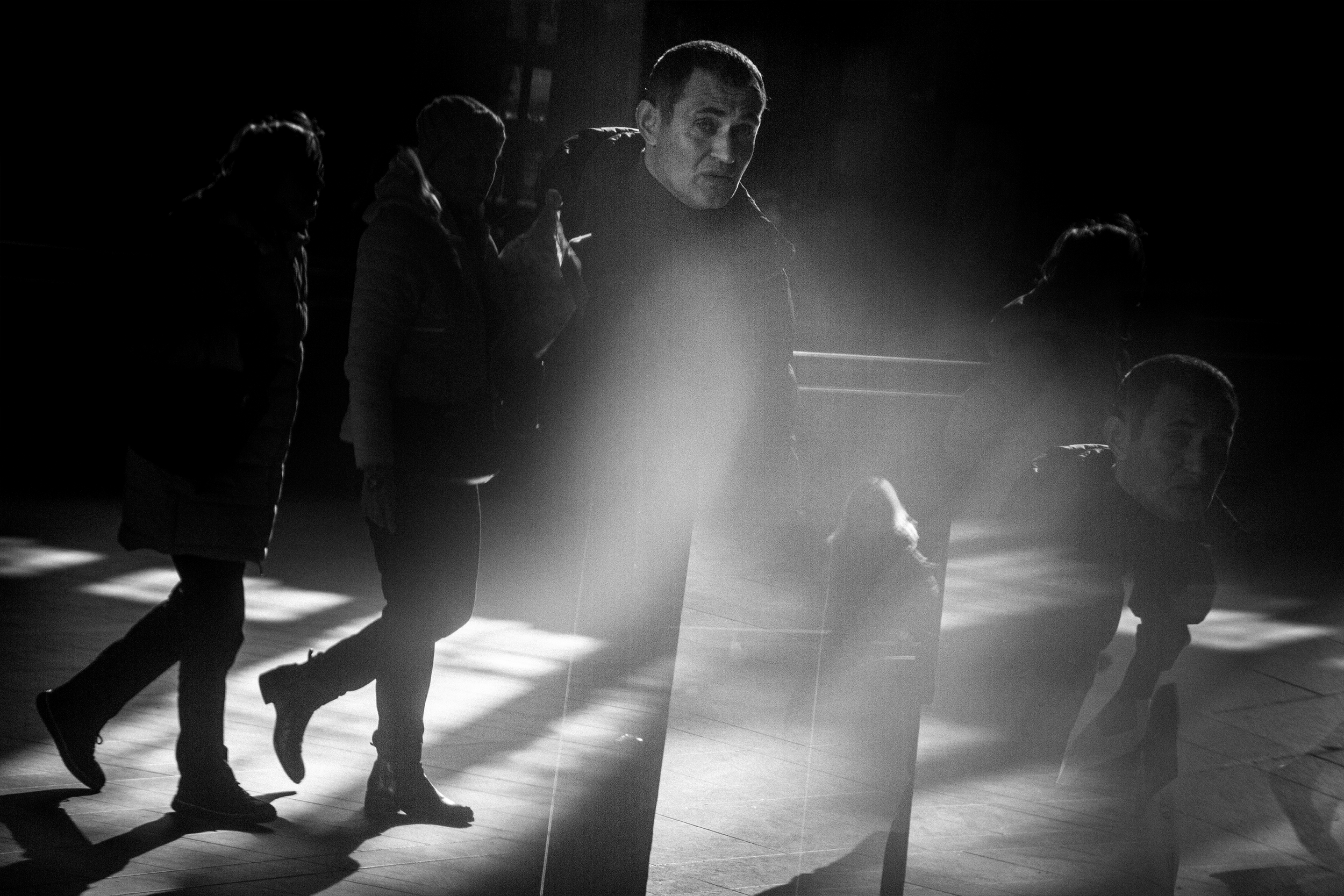 A black and white photo showing multiple people in a line, with some walking and one person in focus, illuminated by a light source creating shadows on the ground.