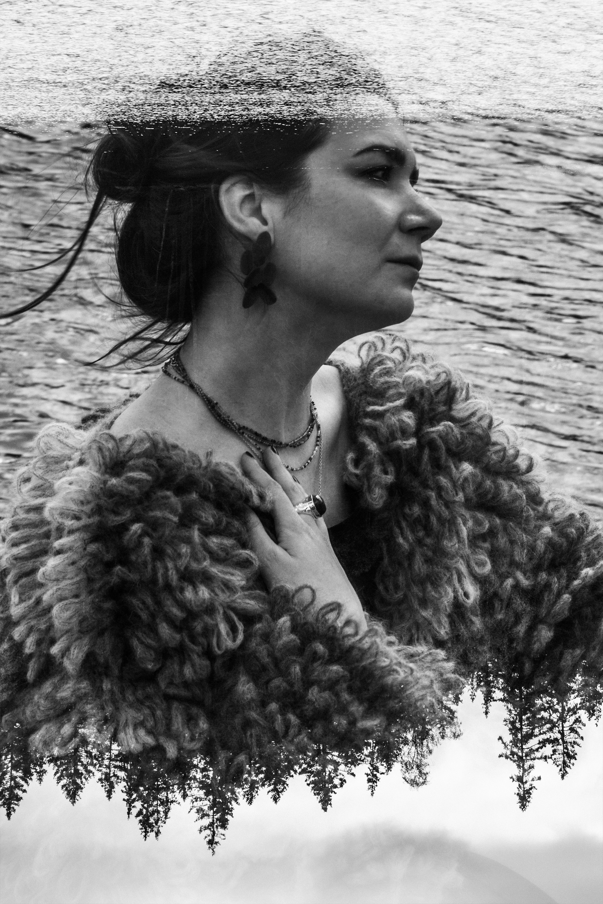 Black and white photo of a woman in profile, wearing earrings and a fuzzy coat, with her hand on her chest, standing in front of a body of water with trees and mountains reflected in it.