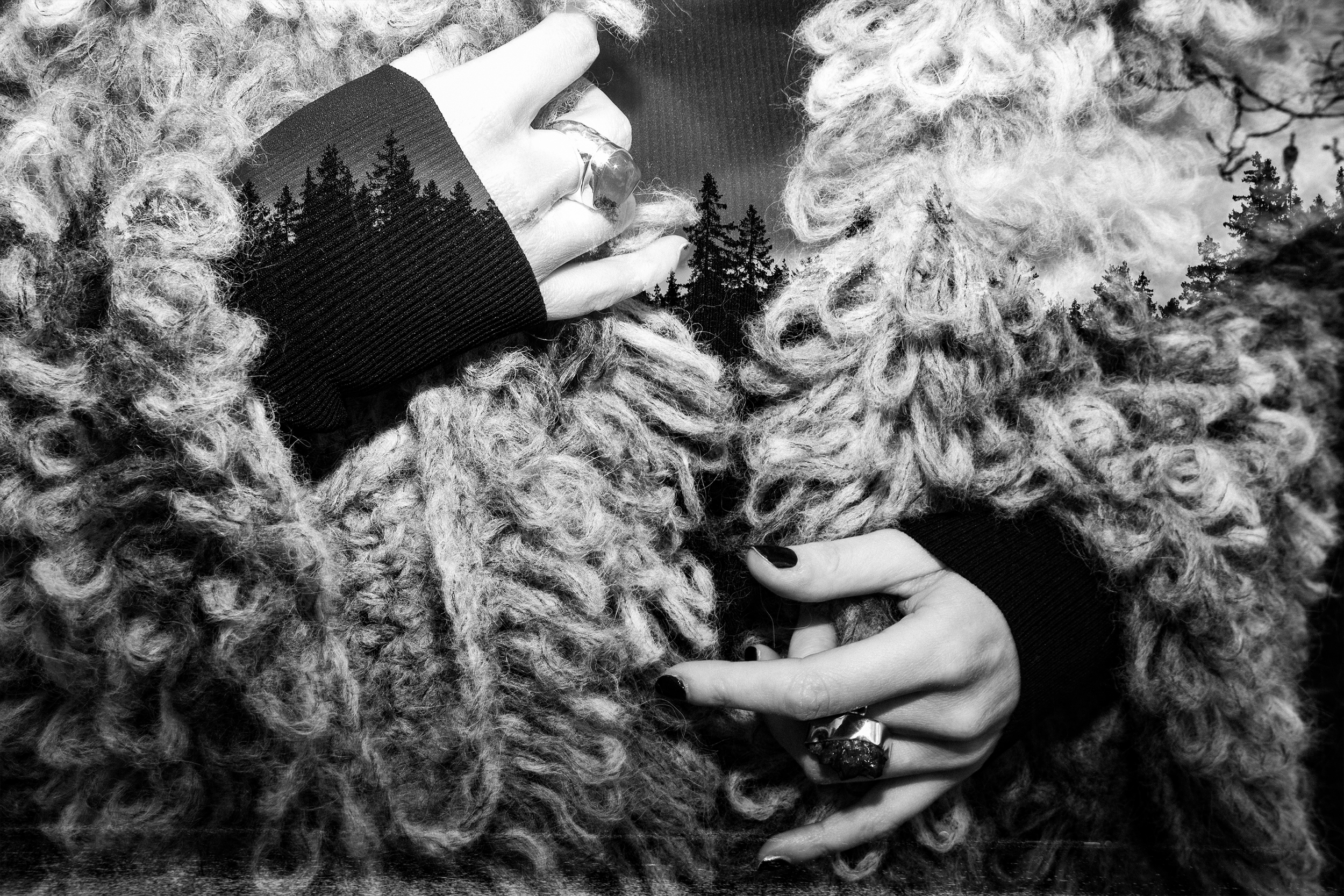 A person with curly hair wearing rings and a black top, holding a fluffy, textured scarf or coat, with a background of trees. The image is in black and white.