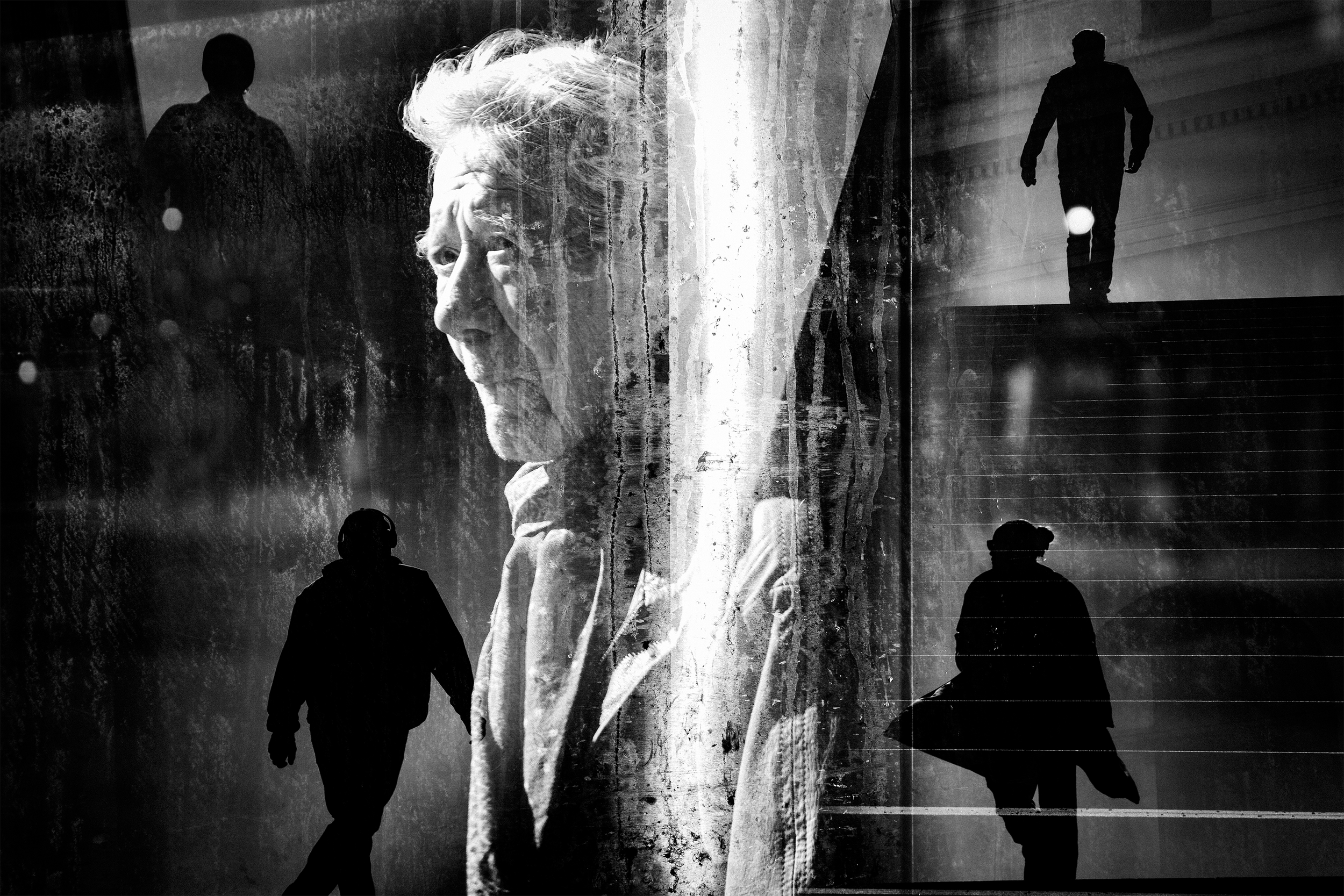 Black and white photo of an elderly woman with a somber expression, layered over silhouettes of three people walking up stairs. The scene appears reflective and introspective. In-camera multiexposure. Remembrance by Oscar Wollsten.