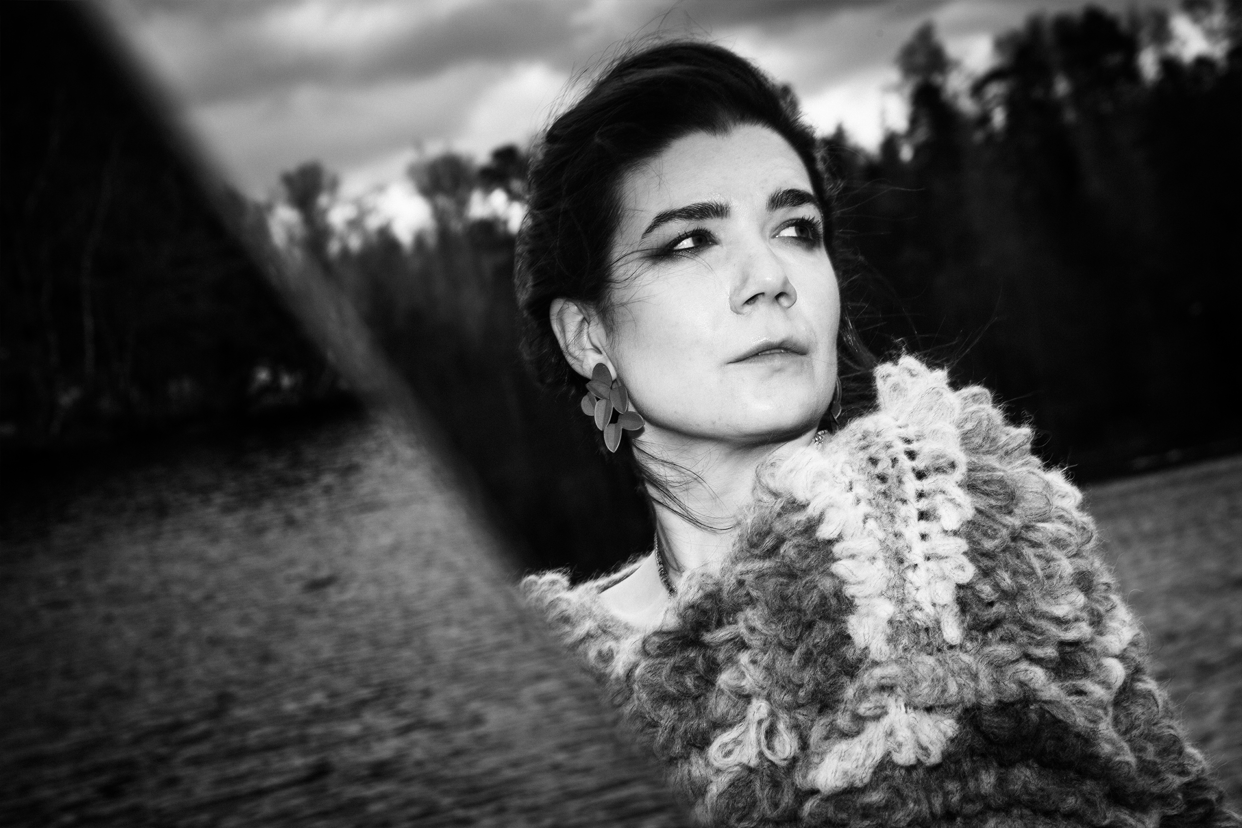 Black and white photo of a woman looking off into the distance, with a blurry landscape in the background. She has dark hair, wears large earrings, and a knitted sweater.
