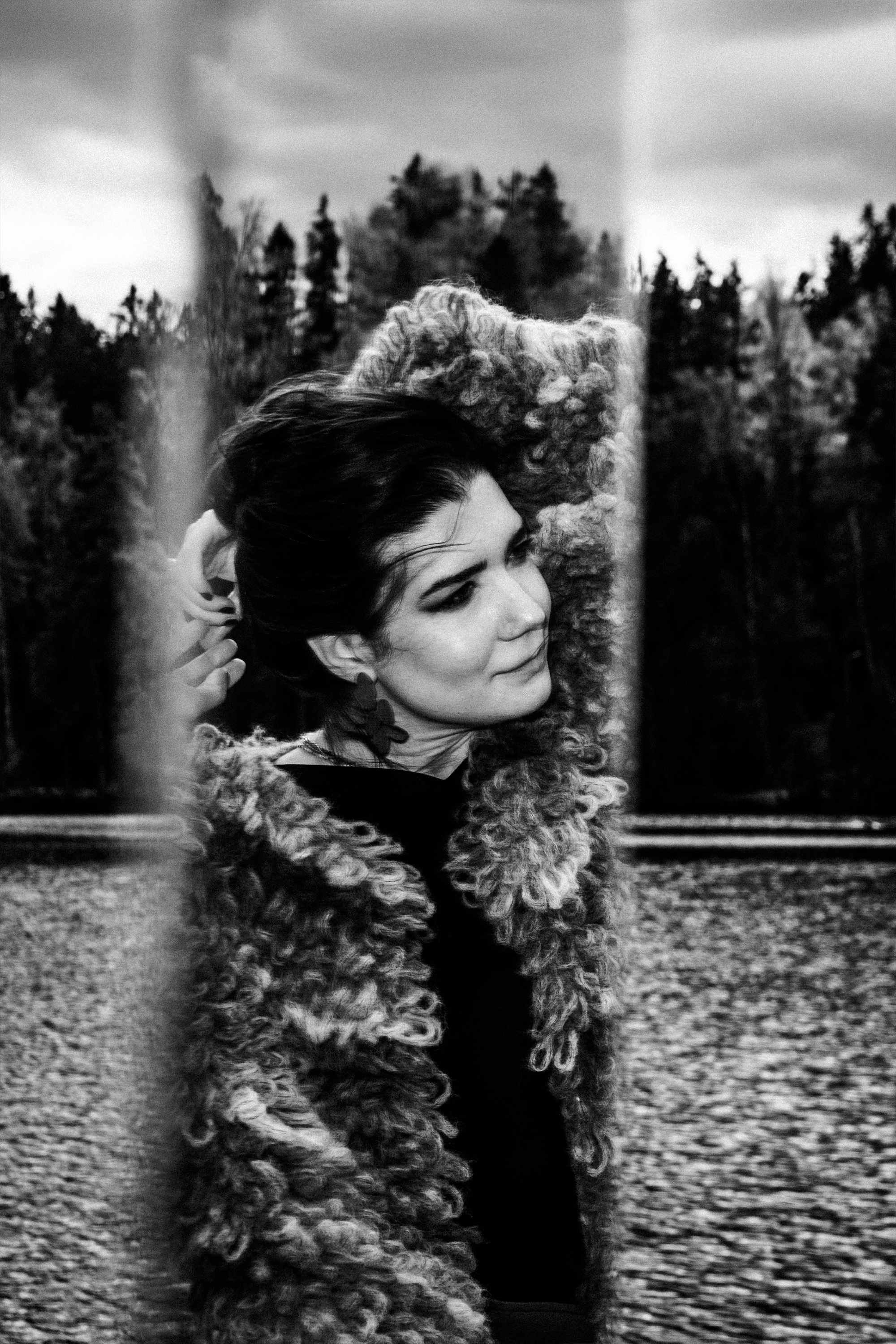 A woman with dark hair is adjusting a curly, thick scarf or coat collar while standing outdoors near train tracks, with a forested area in the background. The photo is black and white.