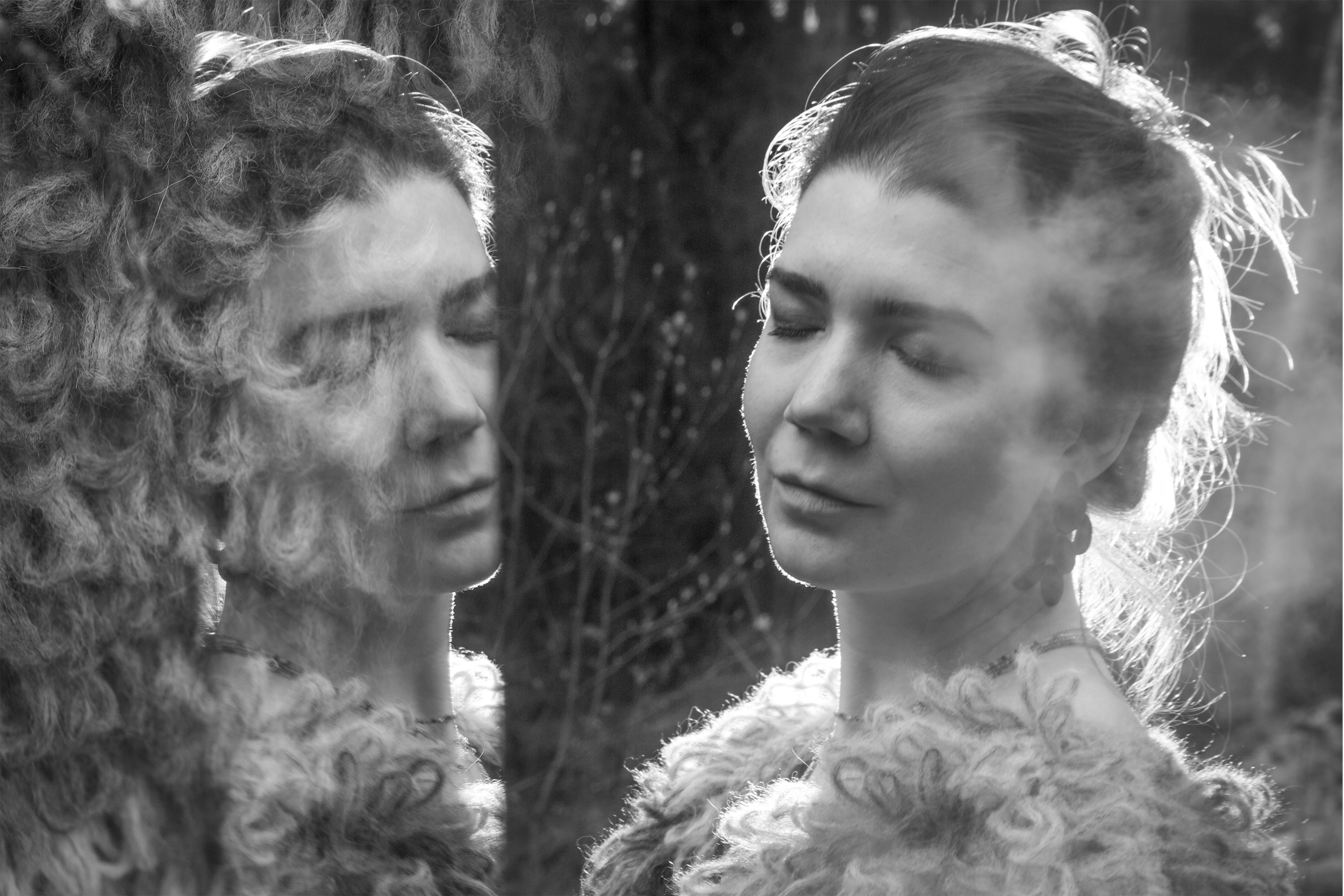 Black and white photo of two women with their eyes closed, facing each other, outdoors with blurred trees in the background.