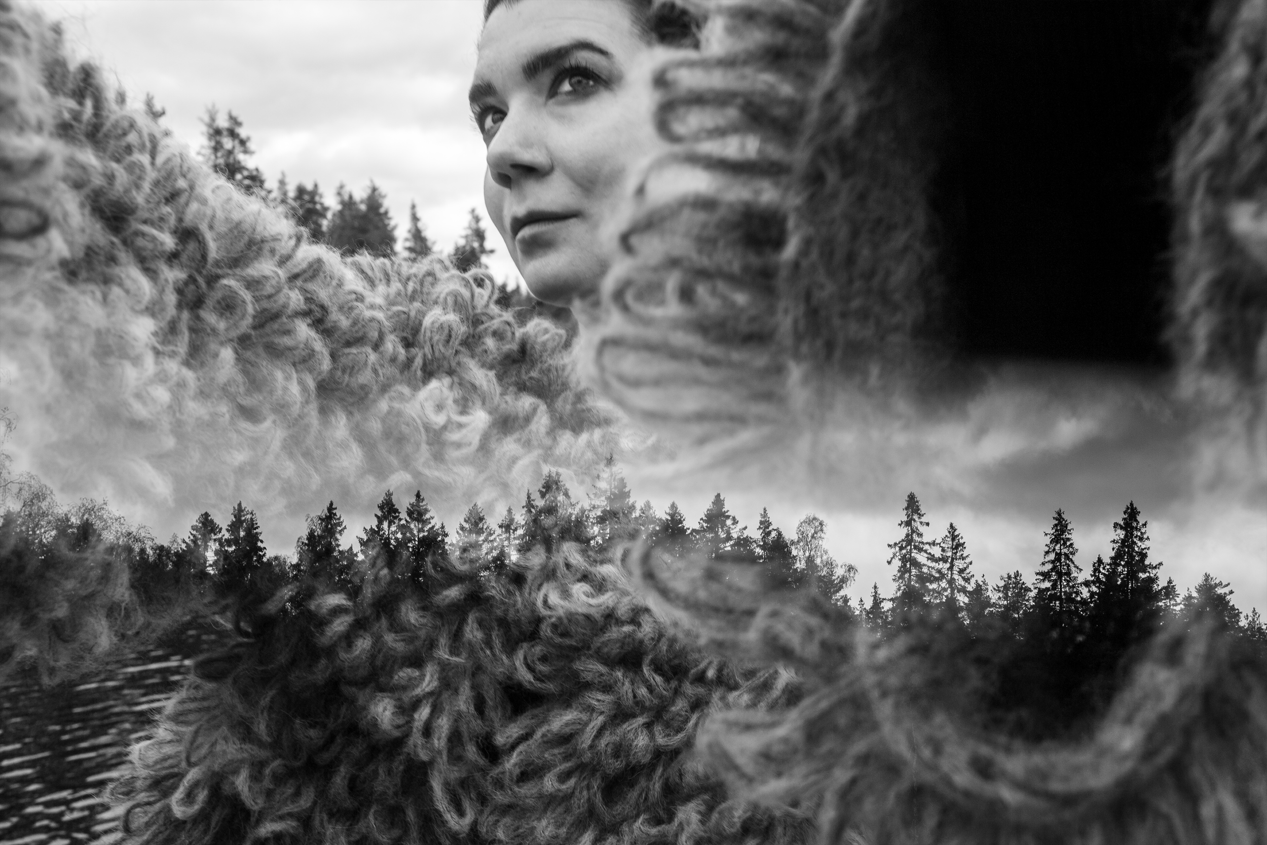 Black and white photo of a woman's face peeking through woolly textiles against a forest background.