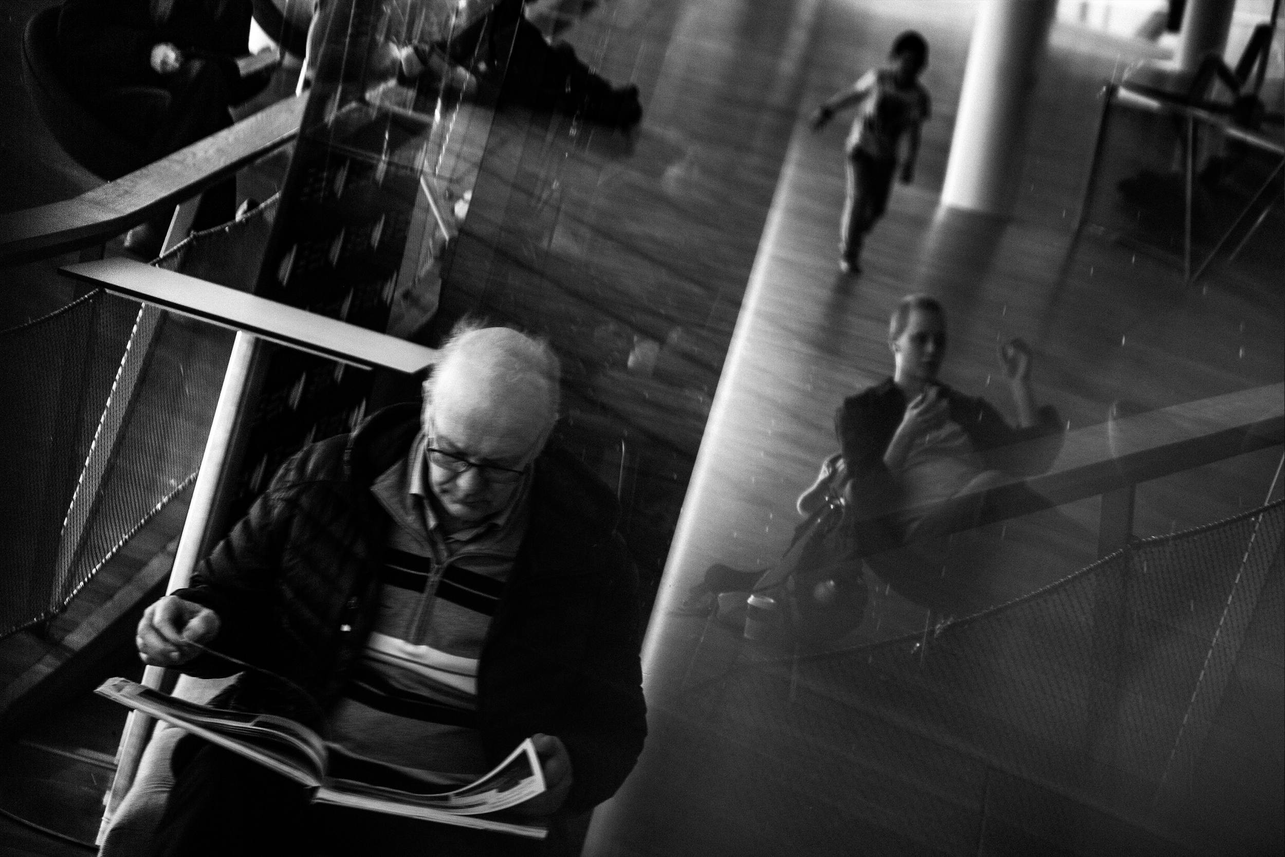 A reflection in a glass wall shows an elderly man with white hair and glasses reading a newspaper, while in the background a young girl is walking, and a woman is seated, all in a modern indoor space with wooden floors.