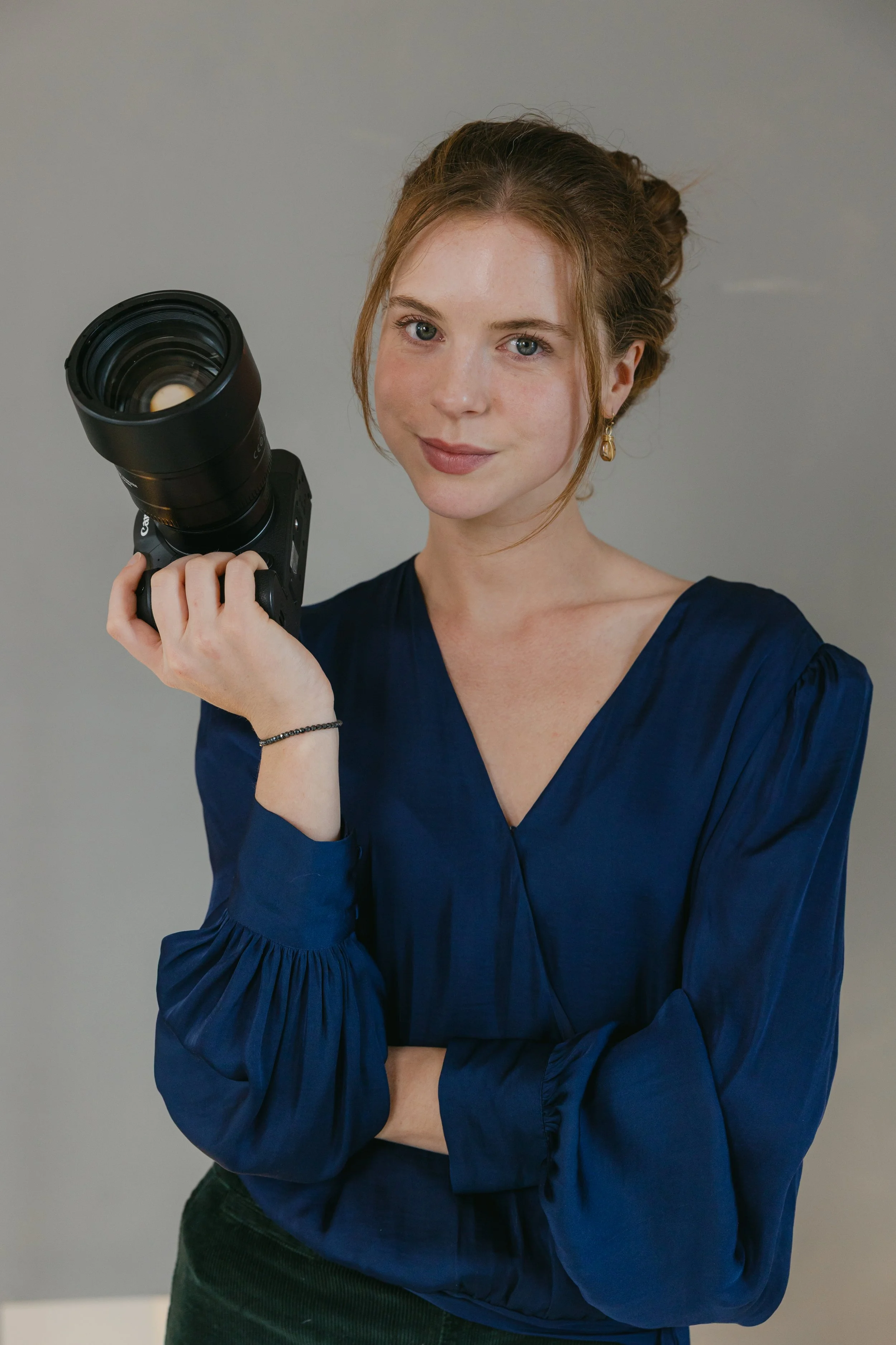 Young woman with red hair in a loose bun, wearing a blue blouse, holding a camera with a large telephoto lens, smiling at the camera.