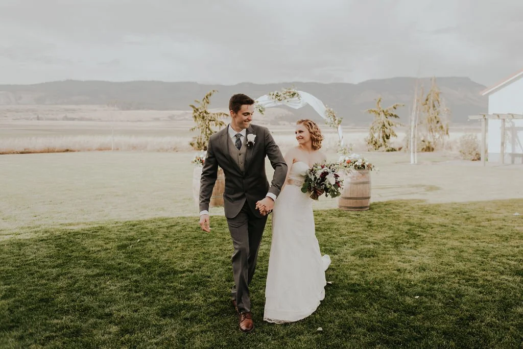 A bride and groom holding hands, walking on a grassy field during a wedding ceremony outdoors, with a scenic landscape in the background.