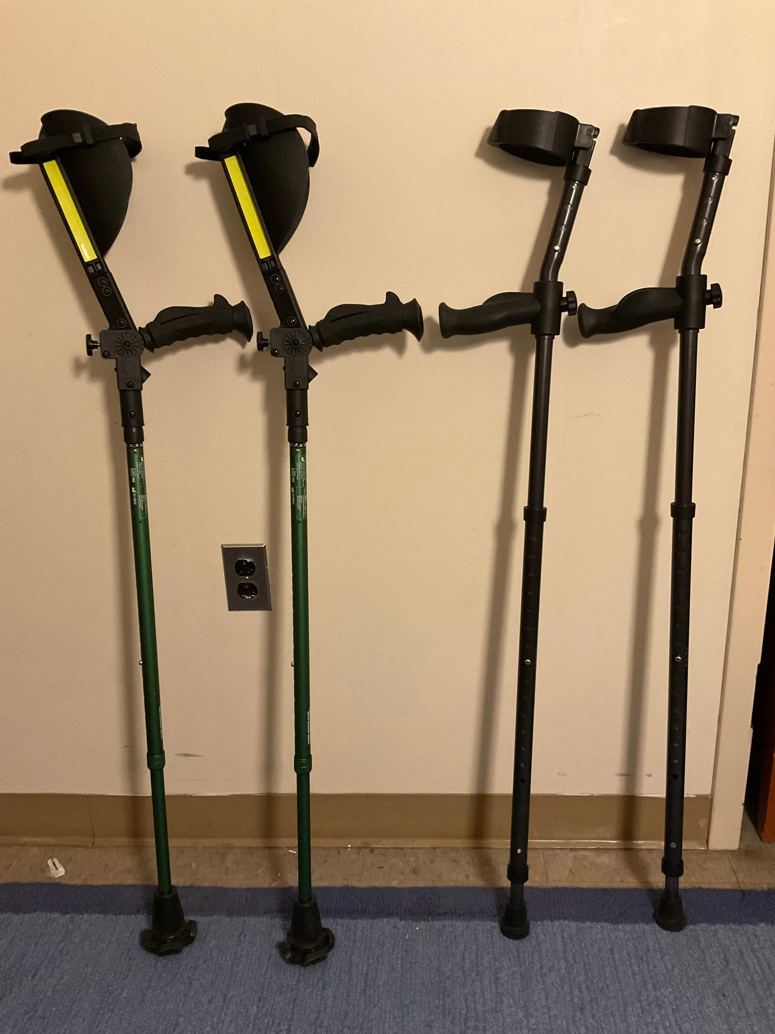 Two pairs of forearm crutches leaning against a wall. The pair on the left are Ergoactives 7G and have large forearm cradles with fluorescent safety strips. The pair on the right are Millennial In-Motion Pros and are all black.