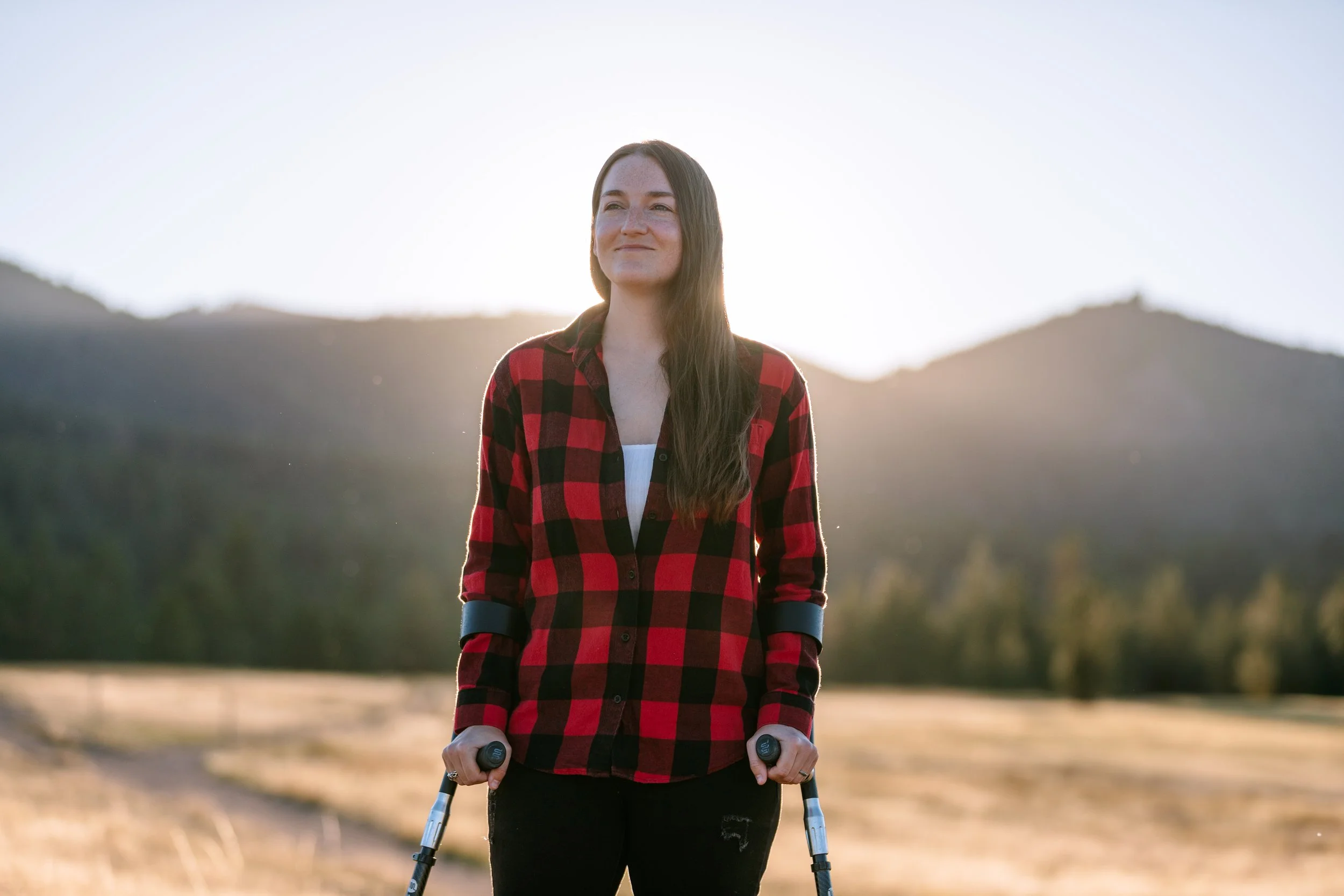 Amy stands with her forearm crutches in front of a mountain scenery, smiling and looking into the distance with the sun glowing behind her.