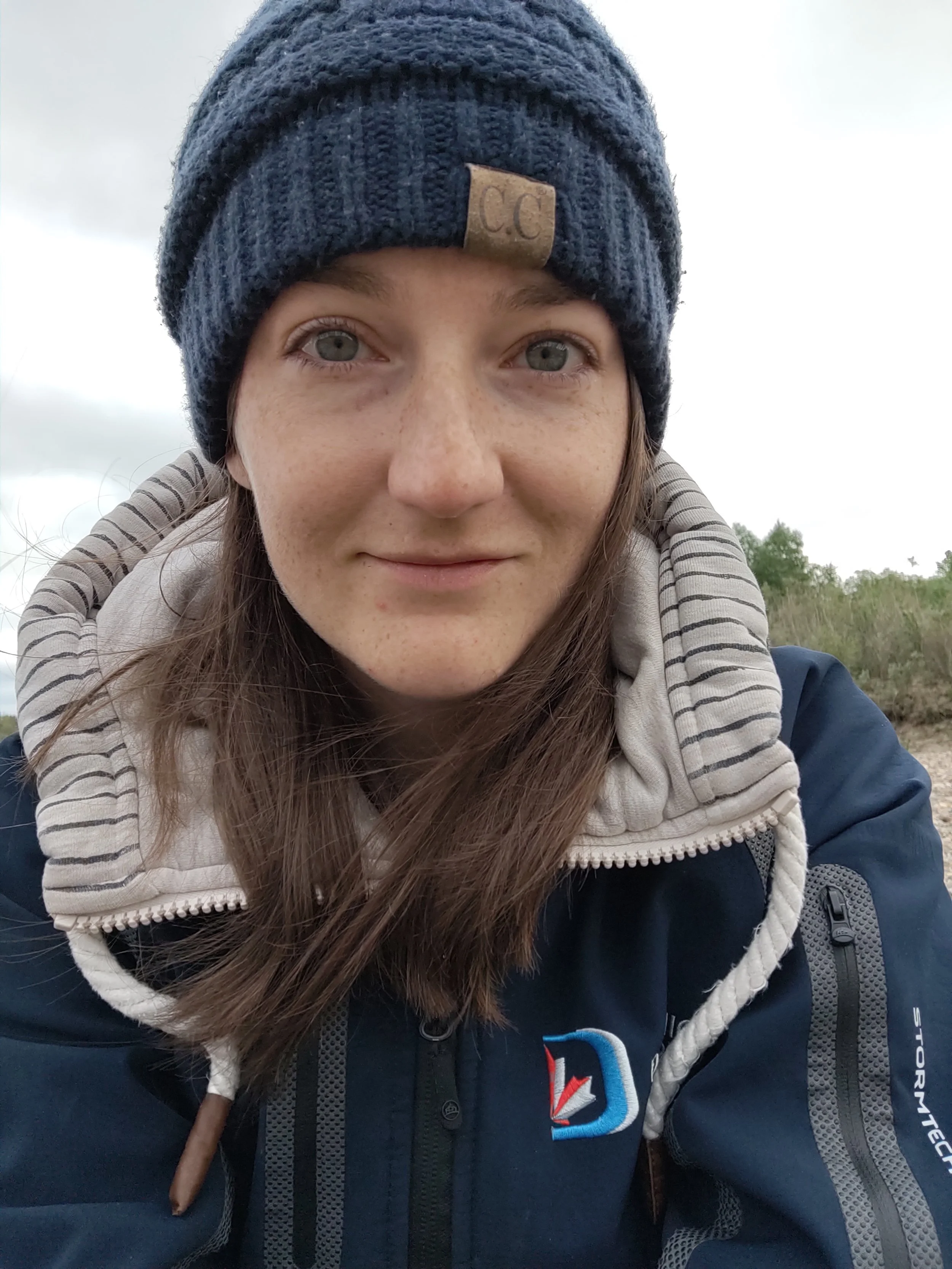Amy grins slightly for a selfie while sitting on a riverbed, her eyes look tired and watery