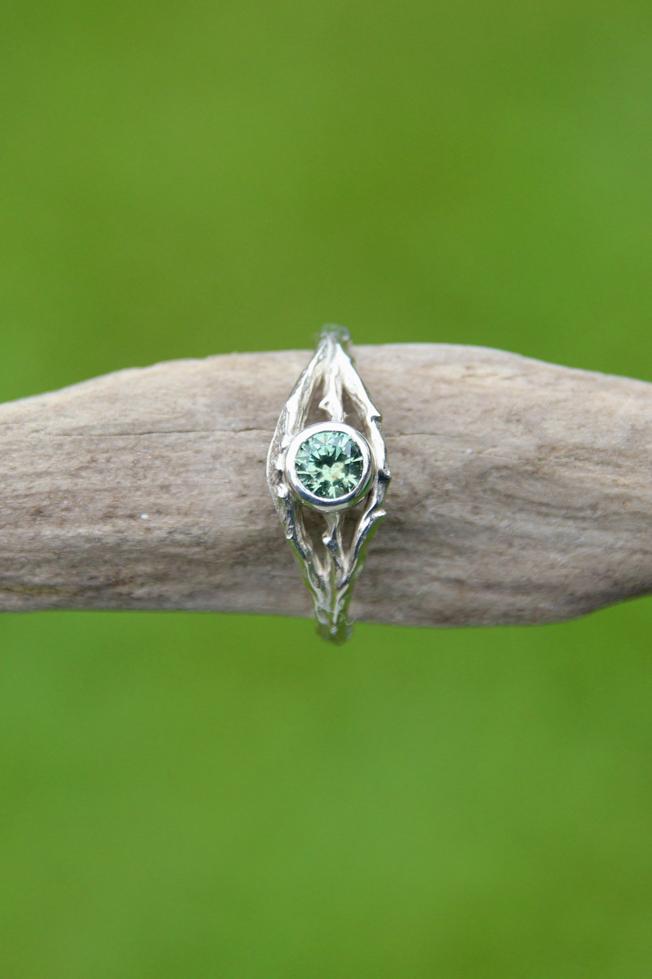 Close-up of a silver ring with a green gemstone, resting on a wooden surface with a blurred green background.