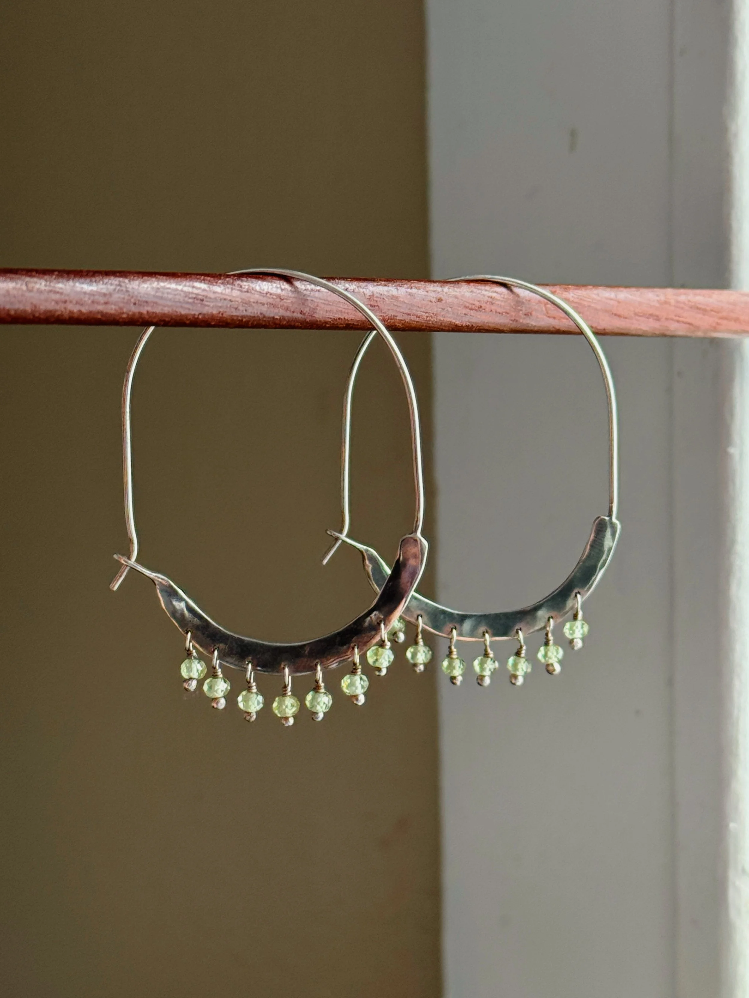 Silver hoop earrings with small green beads hanging from the bottom edge, hanging on a wooden rod.