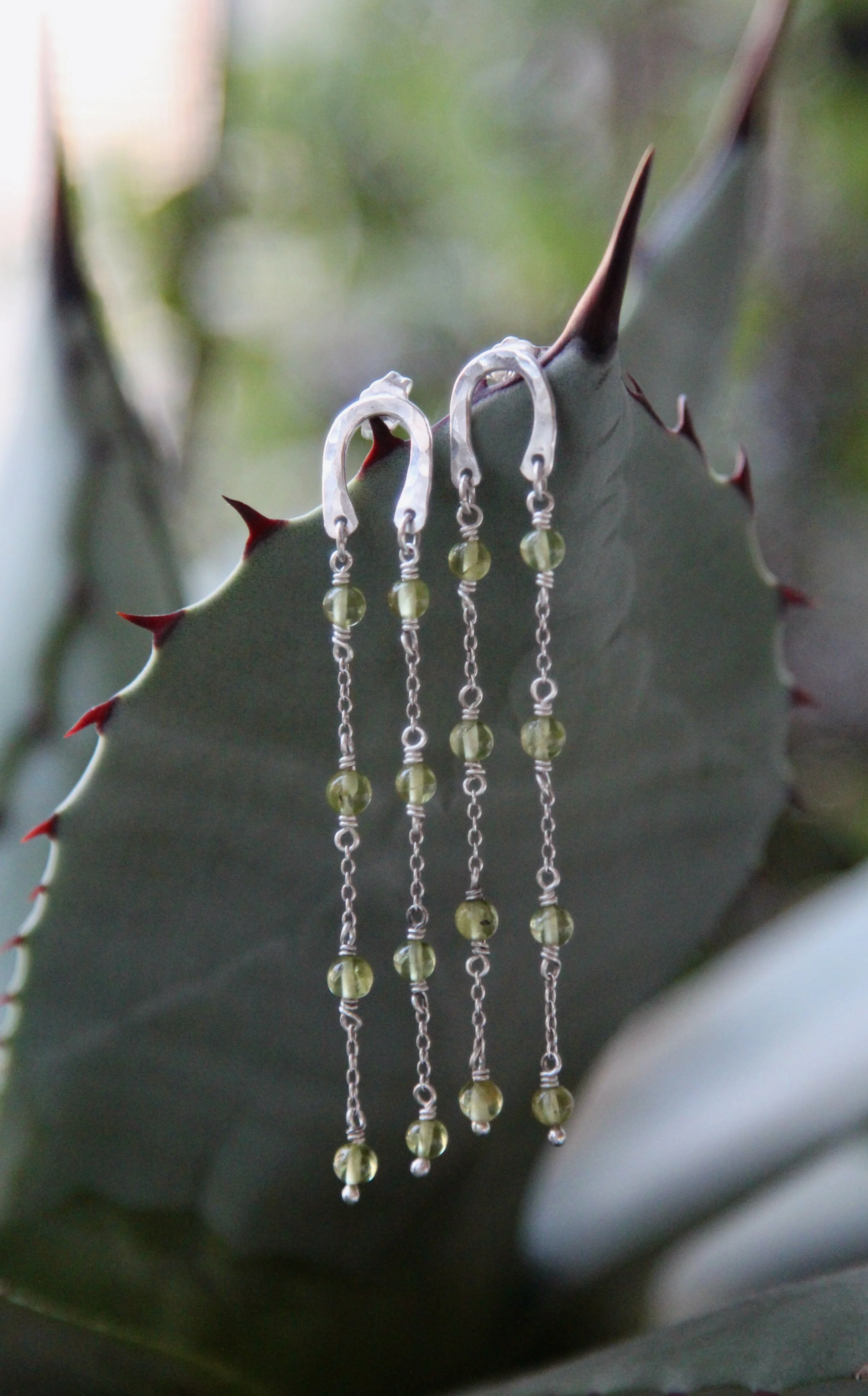 Silver earrings with green beads hanging from a cactus plant with sharp red-tipped thorns.