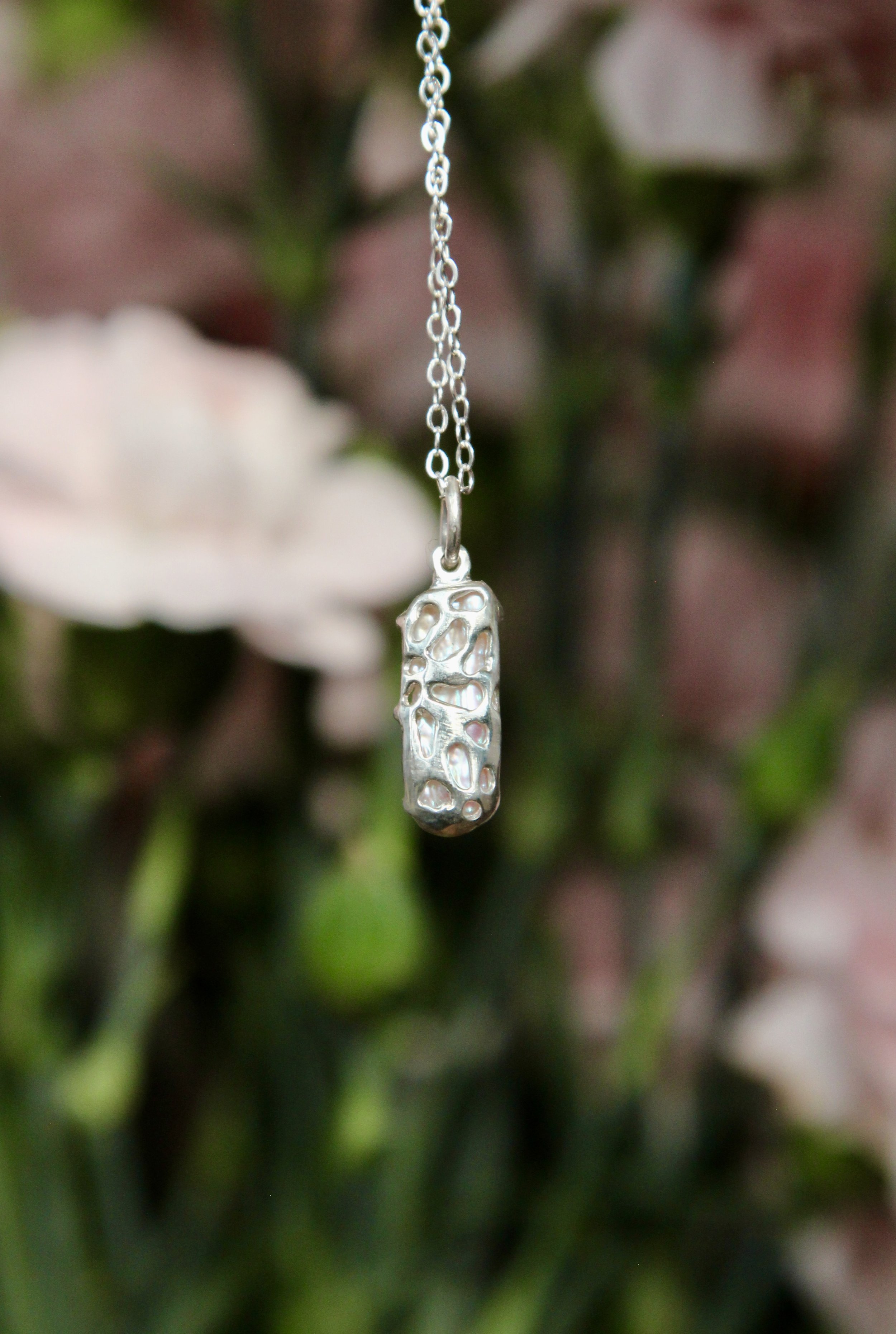 A silver necklace with a pendant featuring irregular, organic-shaped holes, hanging in front of a blurred background of greenery and pinkish flowers.