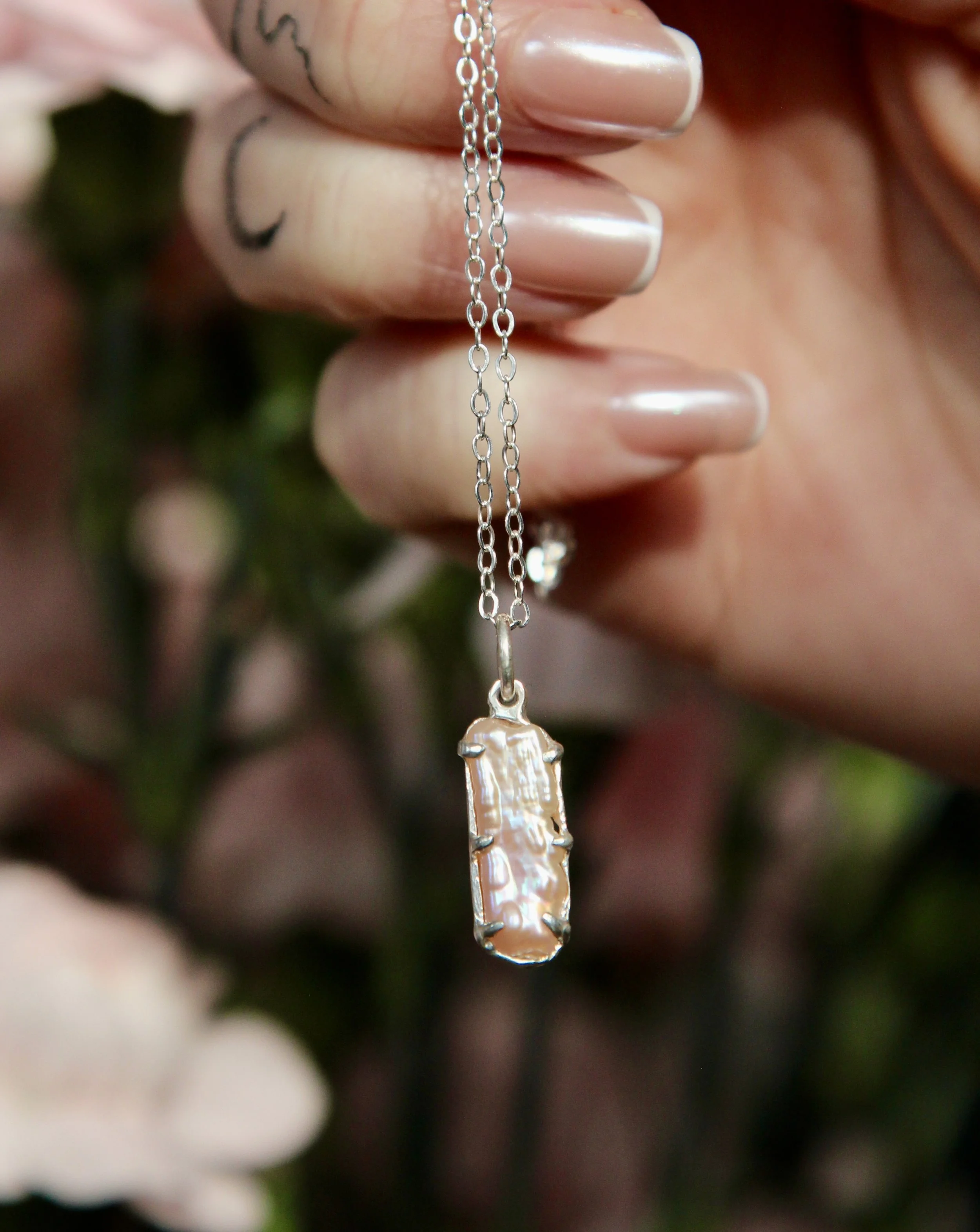 Close-up of a silver chain necklace with a large rectangular pearl pendant, held between fingers, with blurred greenery and flowers in the background.