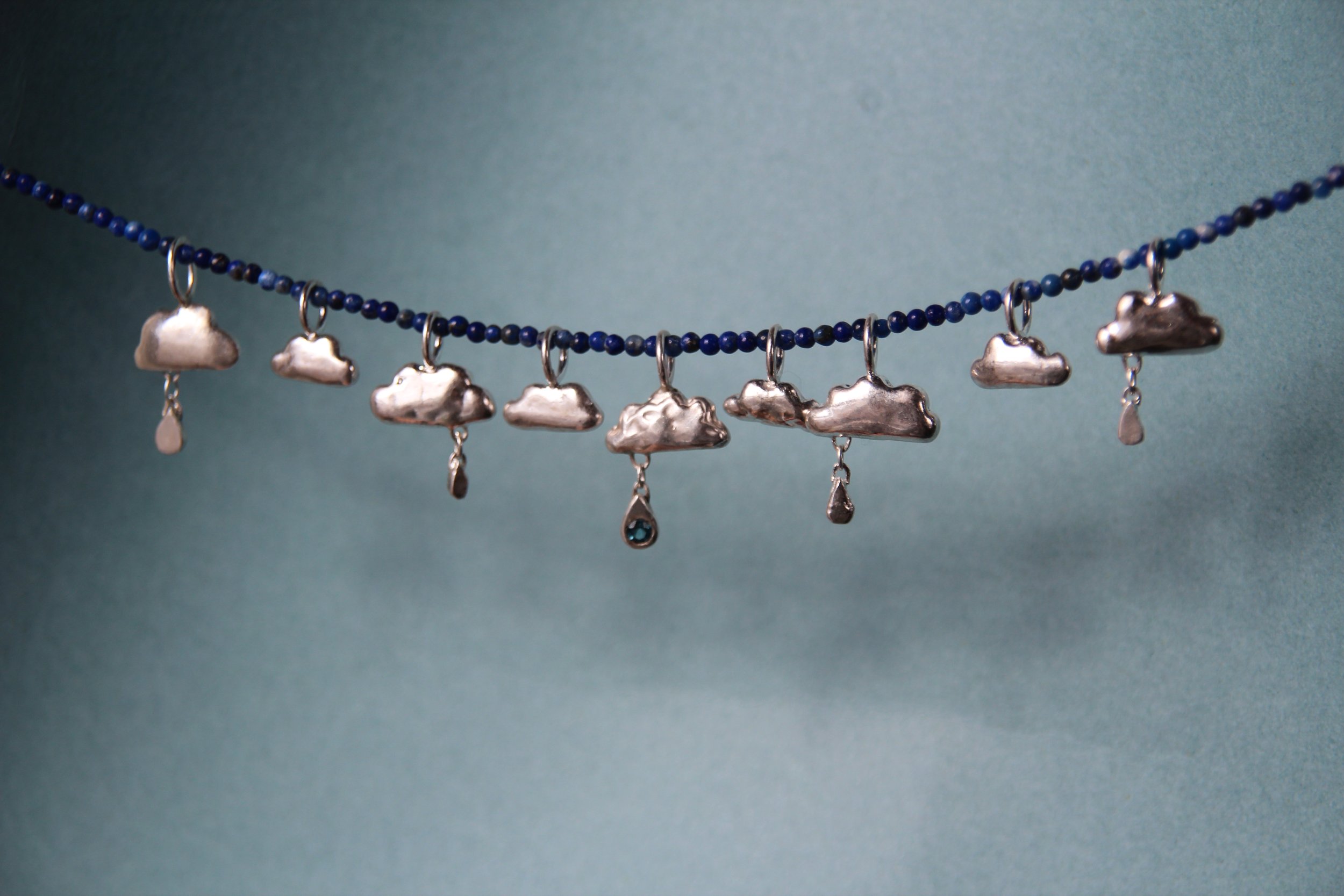 Silver earrings hanging from a blue beaded necklace with cloud-shaped charms and teardrop pendants against a teal background.