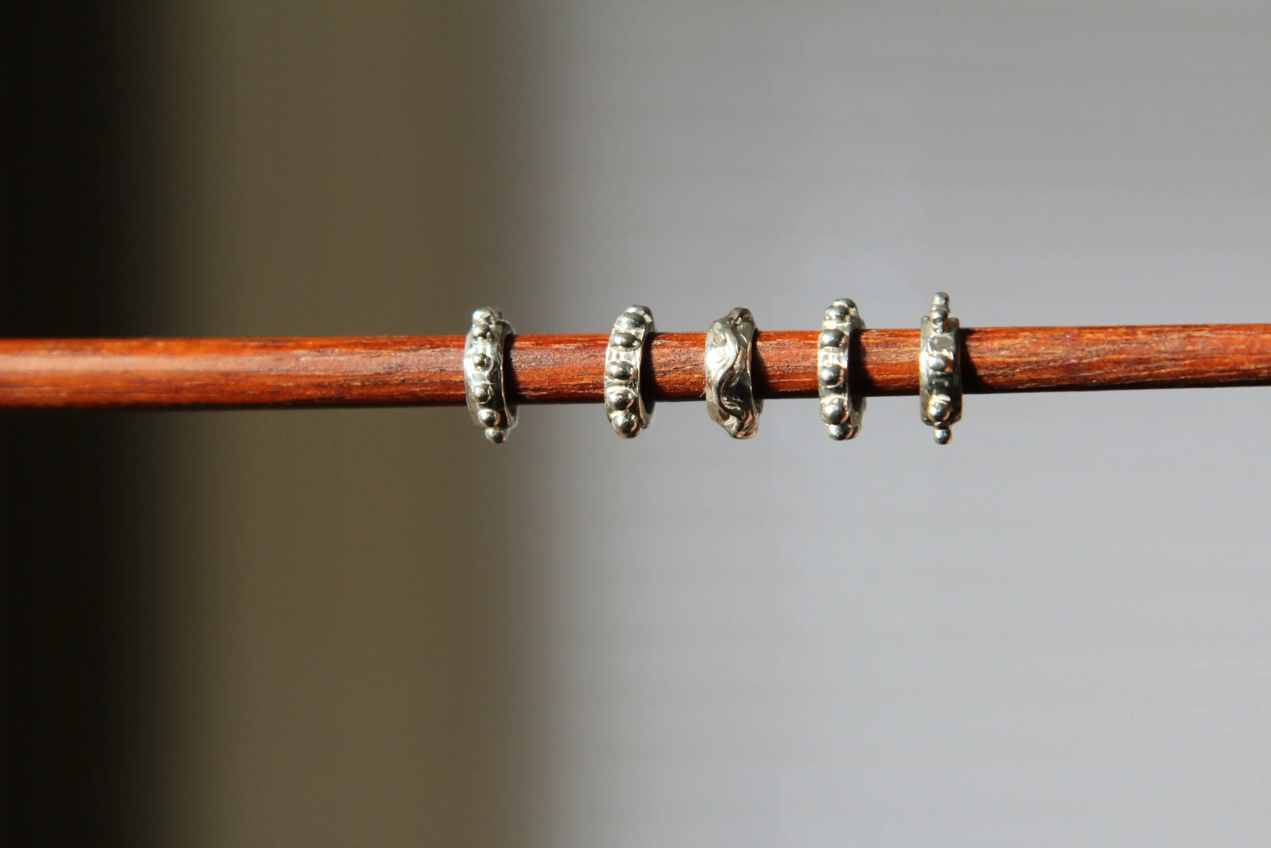 Five silver rings with bead designs hanging on a horizontal wooden stick against a plain background.