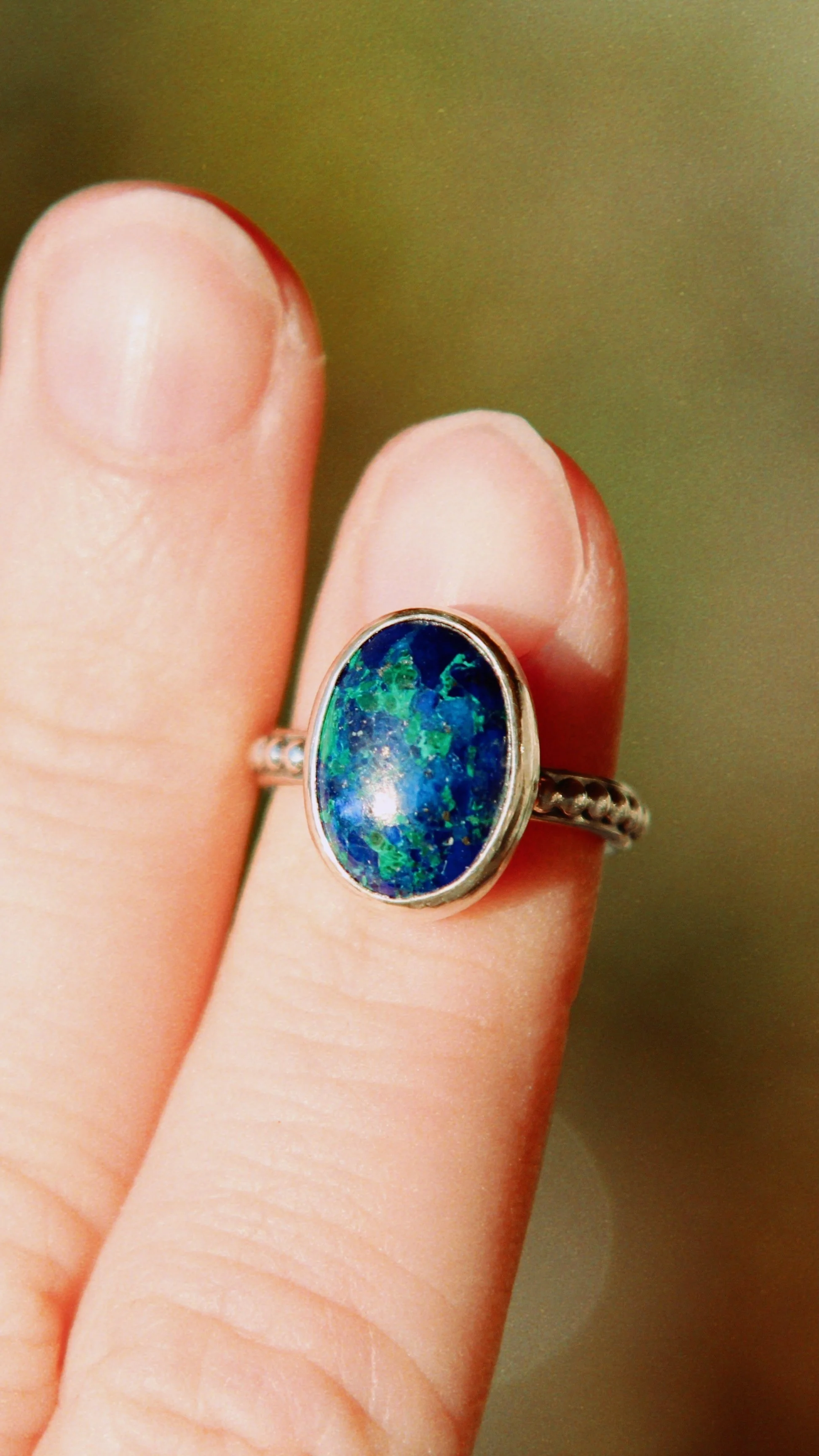 A hand with a silver ring containing an oval-shaped opal gemstone displaying blue and green colors.